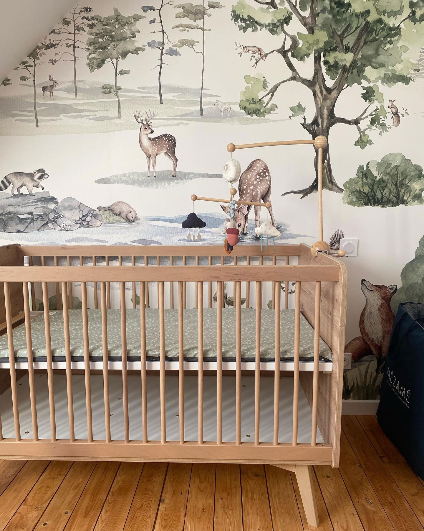 Chambre de bébé avec berceau en bois clair, drap à motifs verts et papier peint avec des animaux de la forêt.
