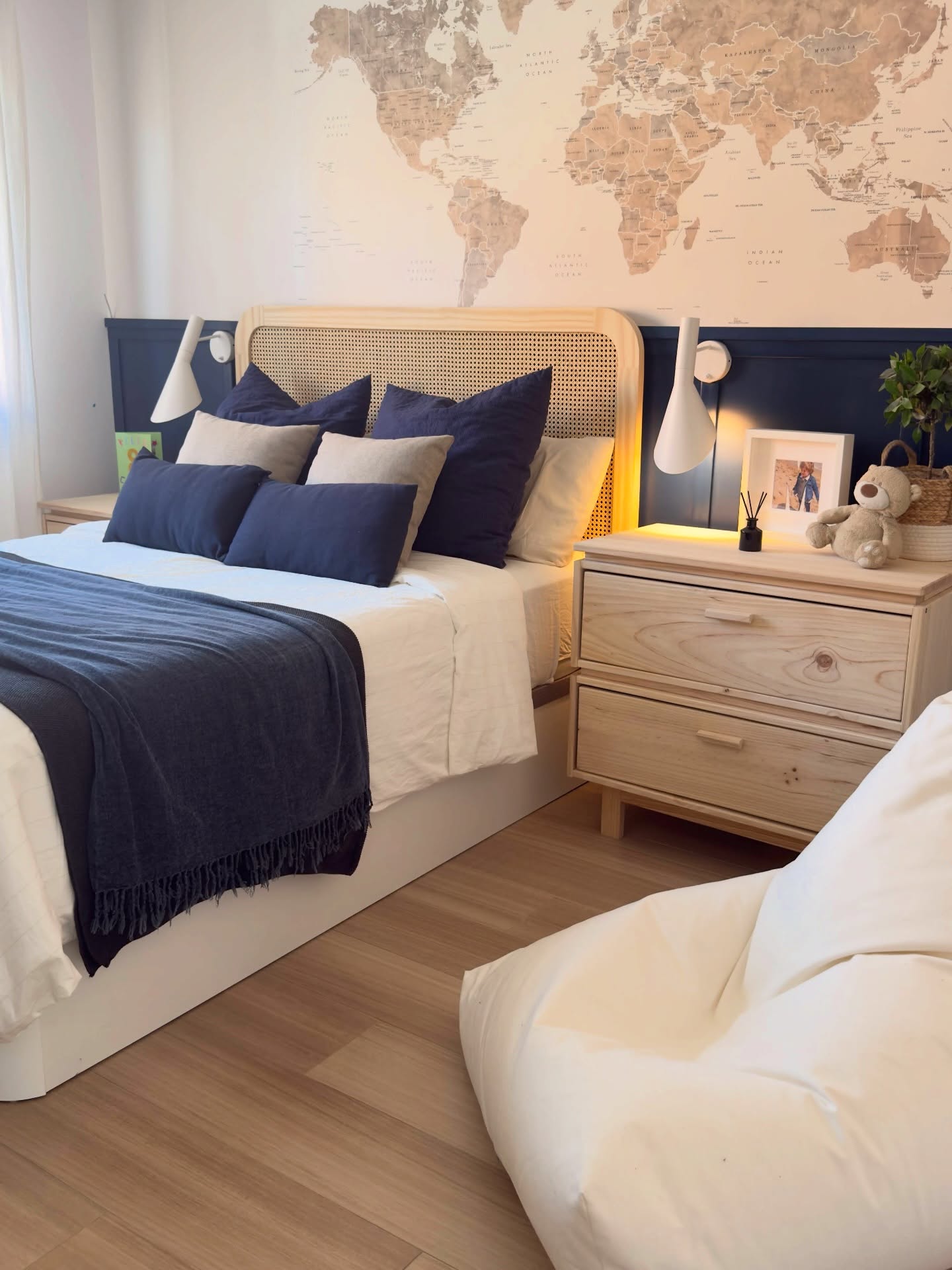 Schlafzimmer mit einer Weltkarten-Wandmalerei, einem Bett mit marineblauen und hellbraunen Kissen und einem Nachttisch aus hellem Holz.