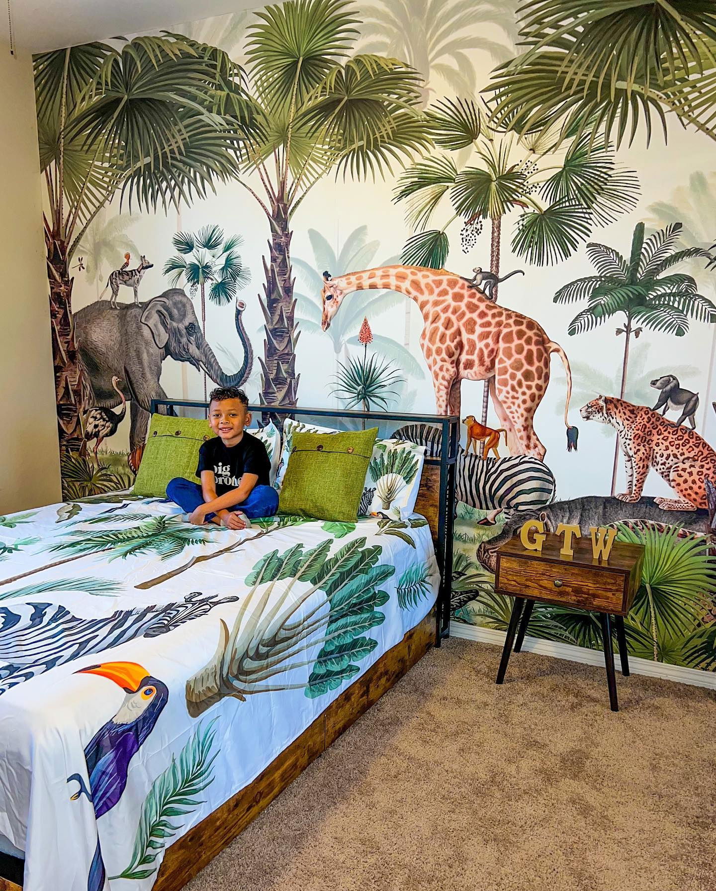 Jongen zit op een bed met jungle-thema beddengoed en een bijpassende jungle muurschildering op de muur.