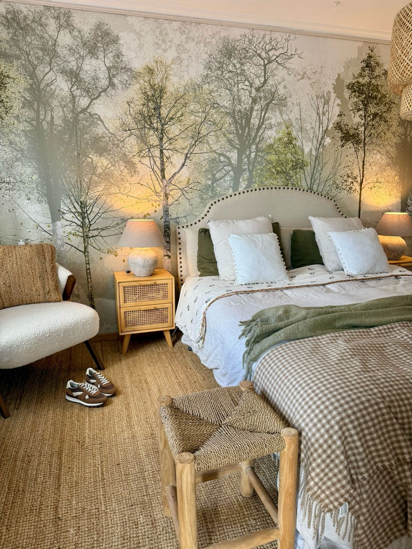 Gemütliches Schlafzimmer mit einer Wandtapete mit Waldmotiv, Polsterbett, Nachttisch aus Rattan, Sessel und einem Hocker aus Webstoff.