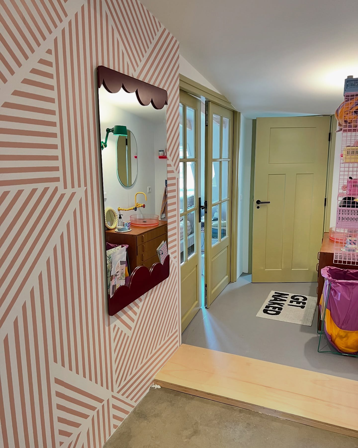 Interior view with a burgundy scalloped mirror on a striped wall, reflecting a bathroom. A "Get Naked" bathmat is visible in the hallway.