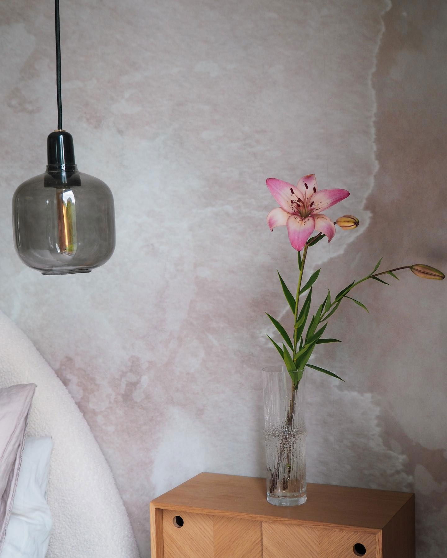 Escena de dormitorio con un lirio rosa en un jarrón sobre una mesita de noche de madera, una lámpara colgante de cristal gris y papel tapiz texturizado.