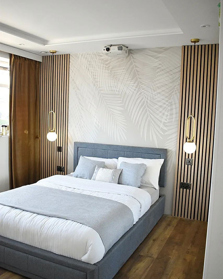Bedroom with gray upholstered bed, white bedding, palm leaf wallpaper, and wood slat accent walls. A projector is mounted on the ceiling.