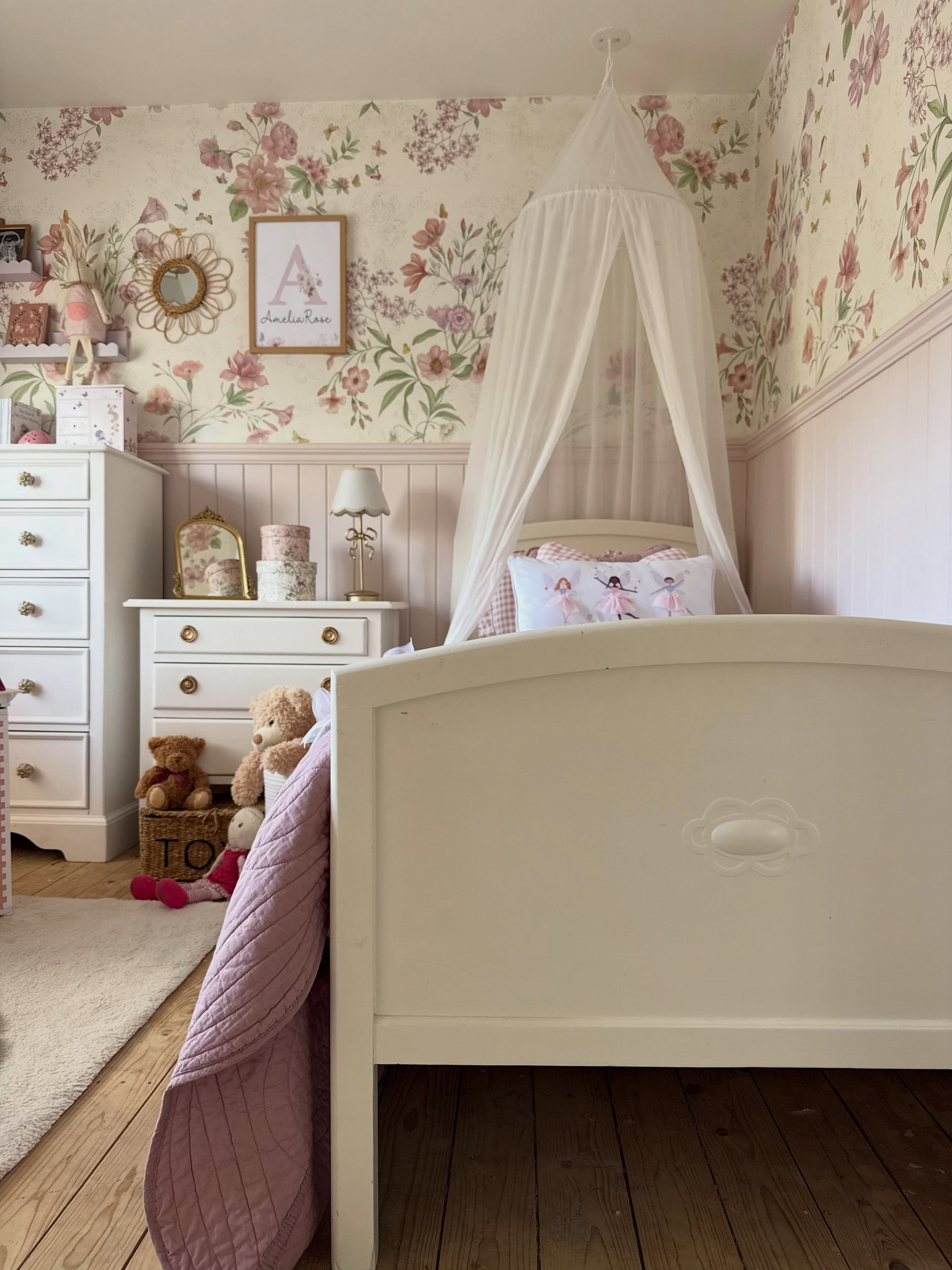 Mädchenzimmer mit floraler Tapete und rosafarbener Wandtäfelung, weißem Himmelbett, weißen Kommoden und einem Ballerina-Kissen. Holzboden.