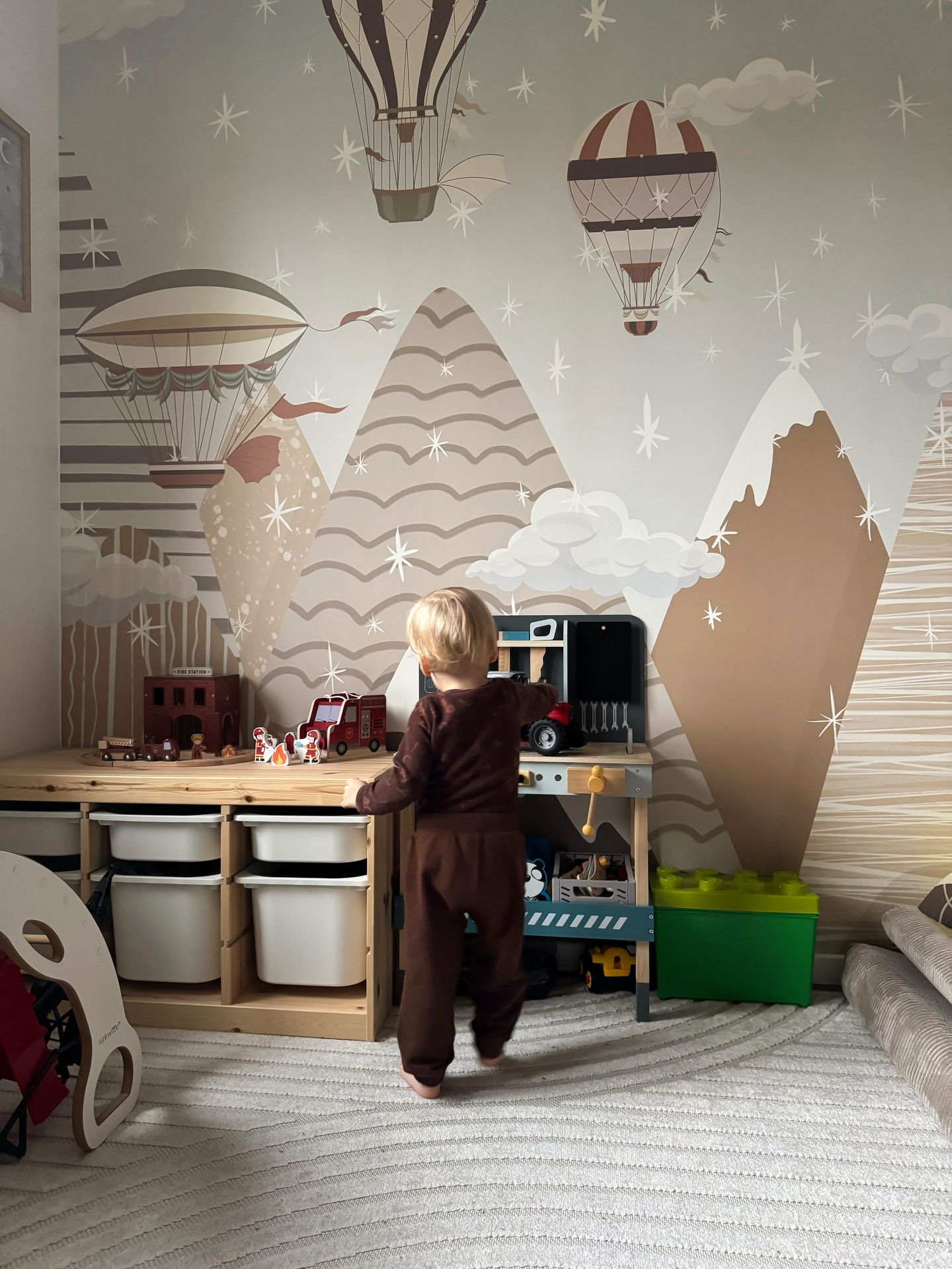 Toddler plays with toys in a room with mountain and hot air balloon wallpaper.