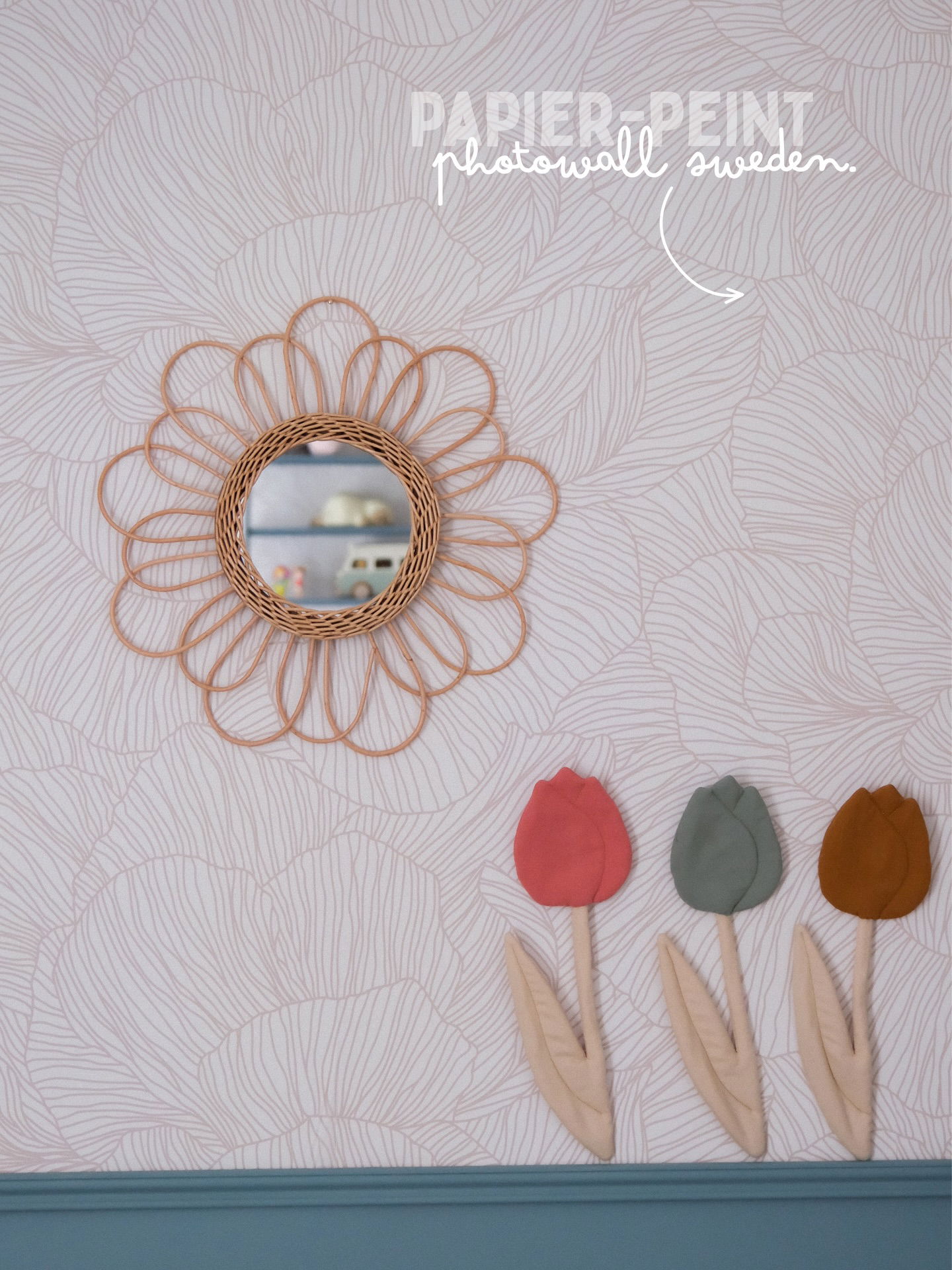 A wicker flower mirror hangs on wallpaper above three fabric tulips in coral, sage, and rust.