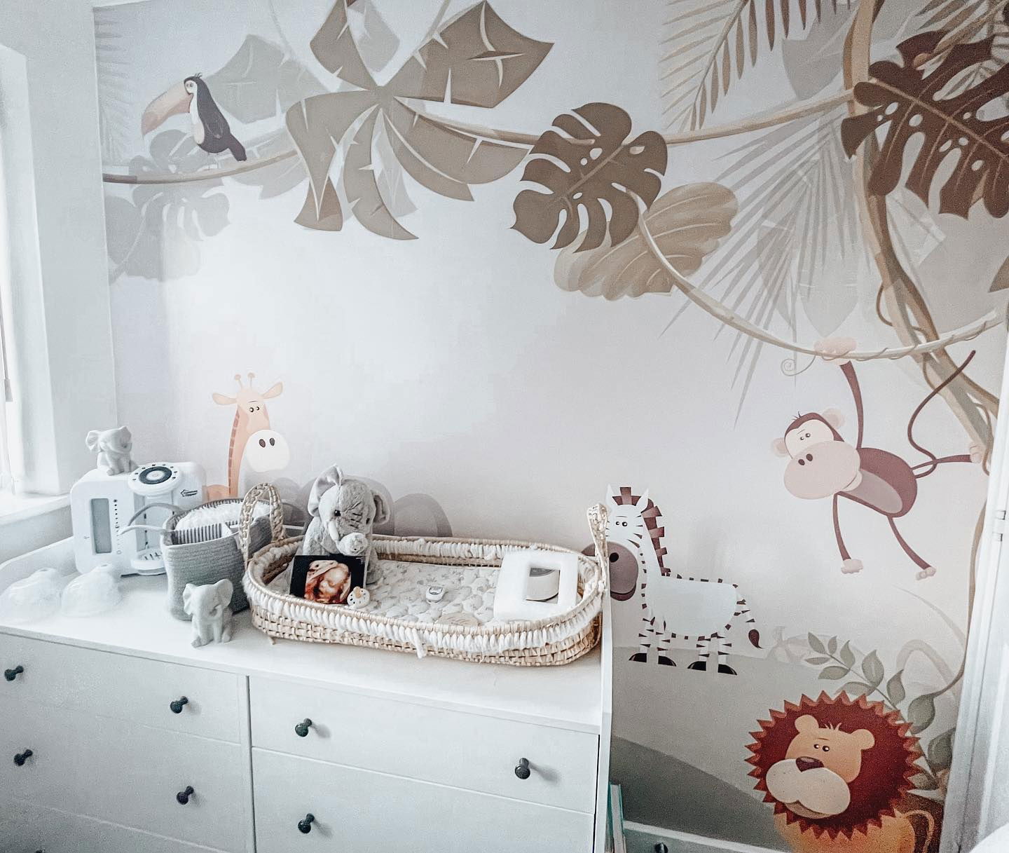 Chambre de bébé avec décorations murales sur le thème de la jungle, commode blanche et panier à langer en osier.