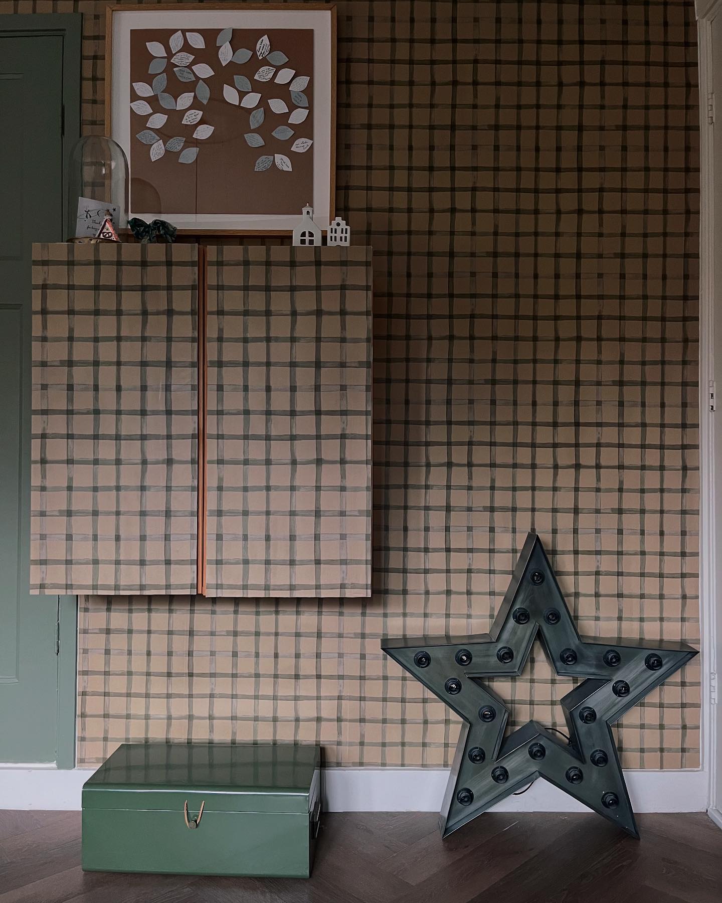 Interior with plaid wallpaper, cabinet, framed leaf art, green box, and star-shaped light.