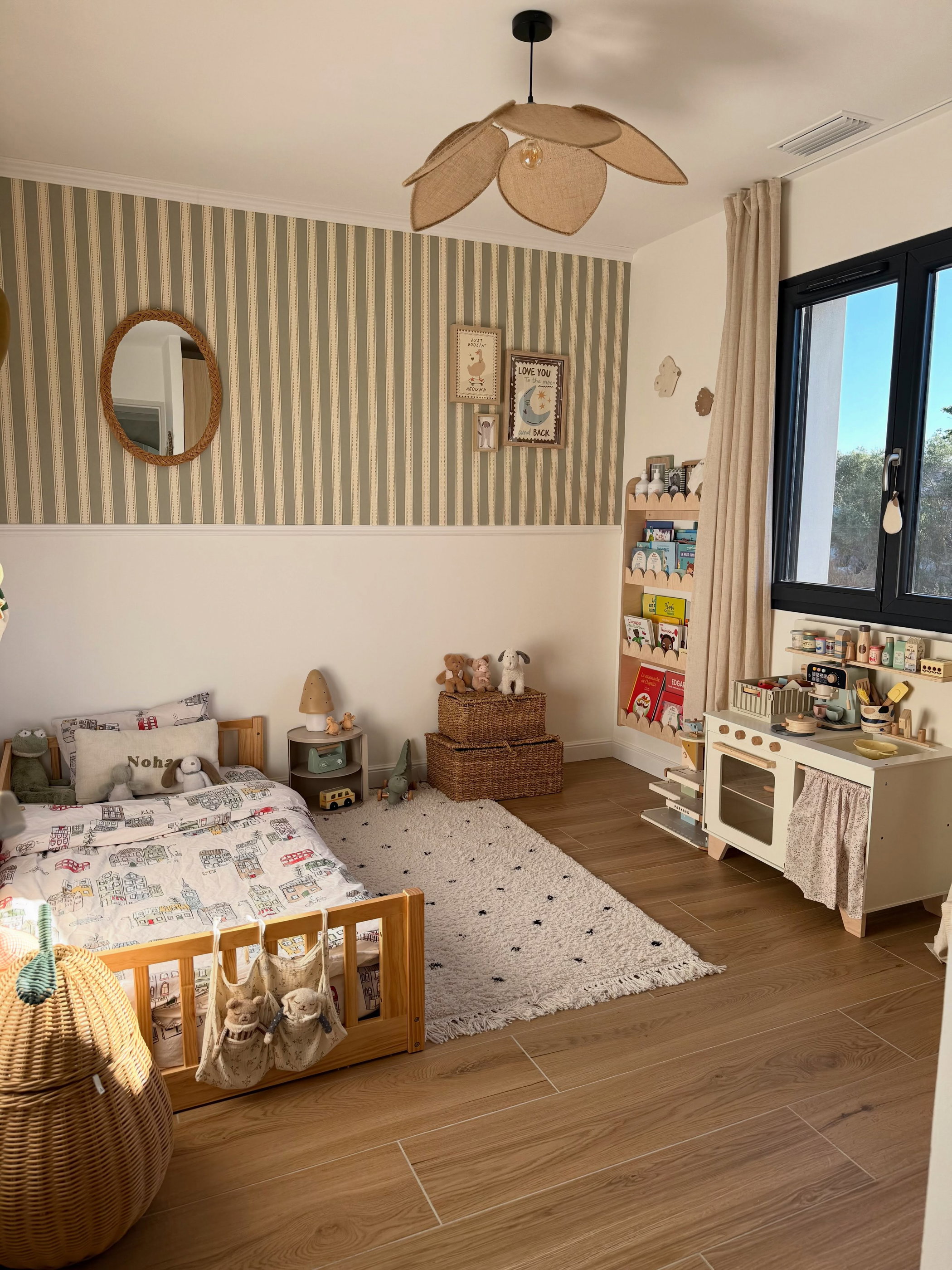 A child's bedroom with a wooden bed, a white play kitchen, and a cream rug with black dots. Walls feature green and white striped wallpaper, and there's a unique leaf-shaped ceiling light.