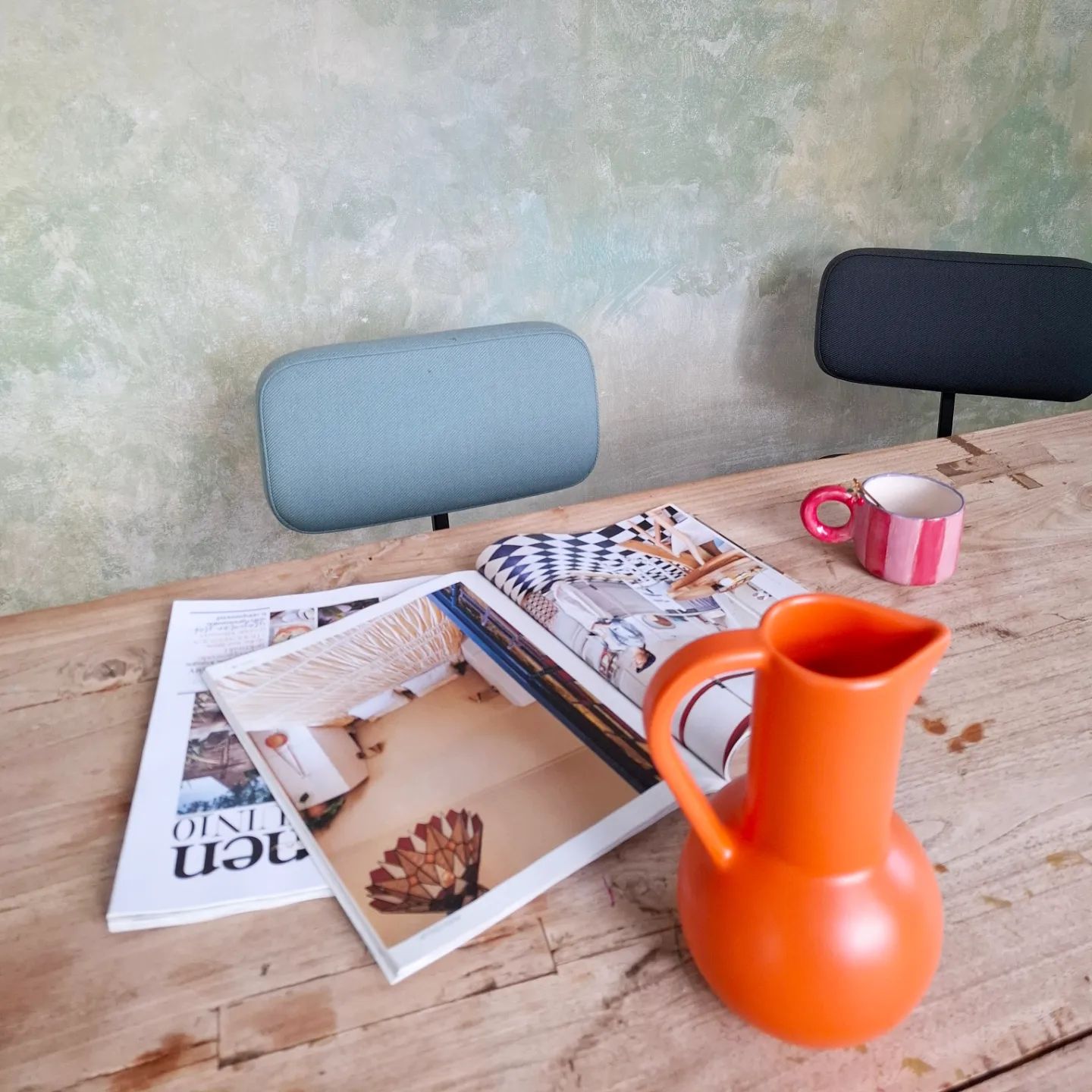 Träbord med tidningar, orange kanna och rosa randig mugg. Två stolar med blått och svart ryggstöd syns.