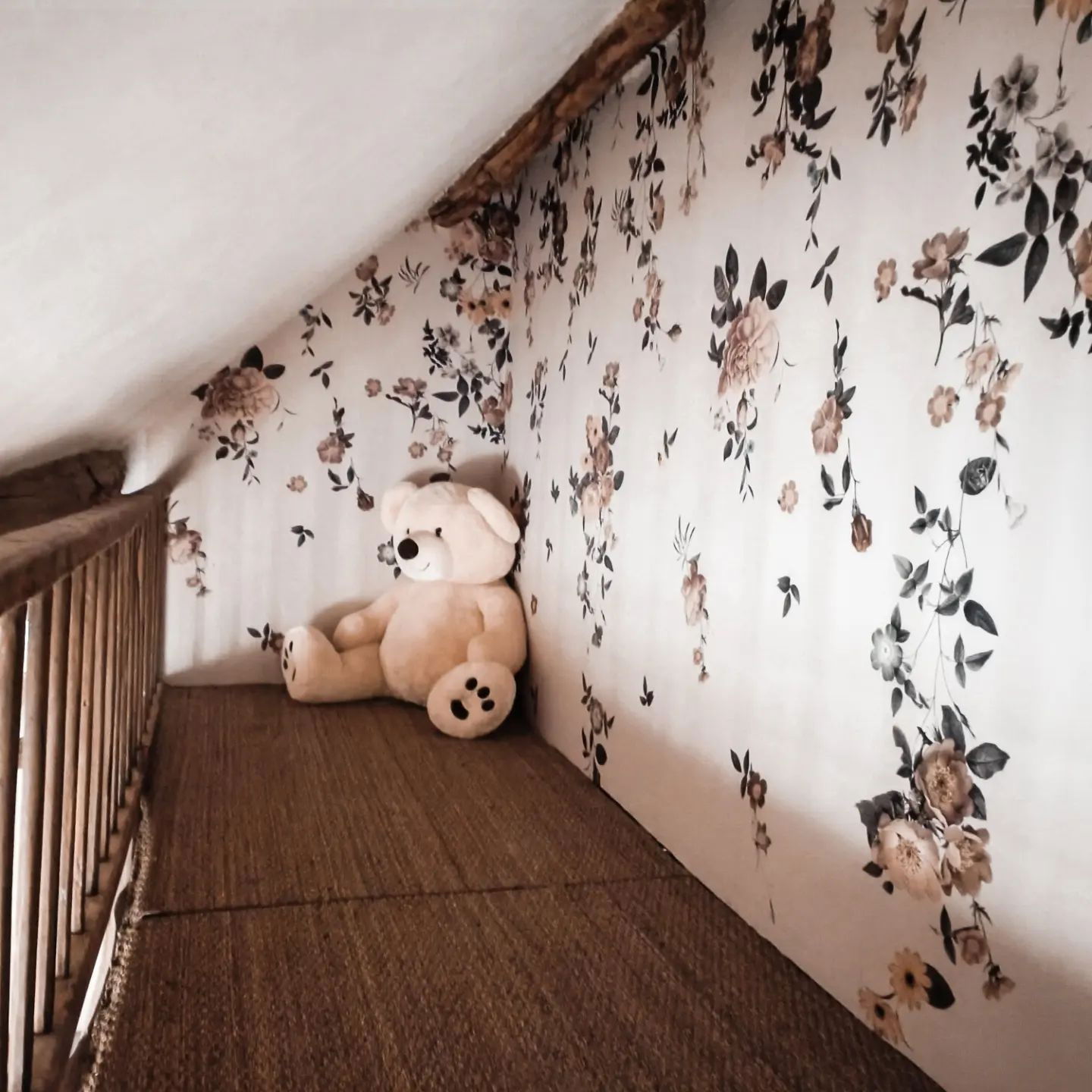 Vindskrymsle med blommiga tapeter, brun vävd matta och en stor, ljusfärgad teddybjörn.