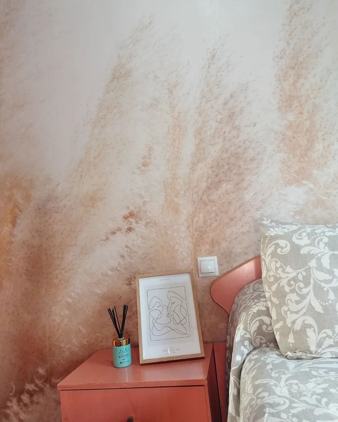 Interior de dormitorio con mesita de noche rosa, arte lineal enmarcado, difusor de caña y ropa de cama estampada contra una pared texturizada.
