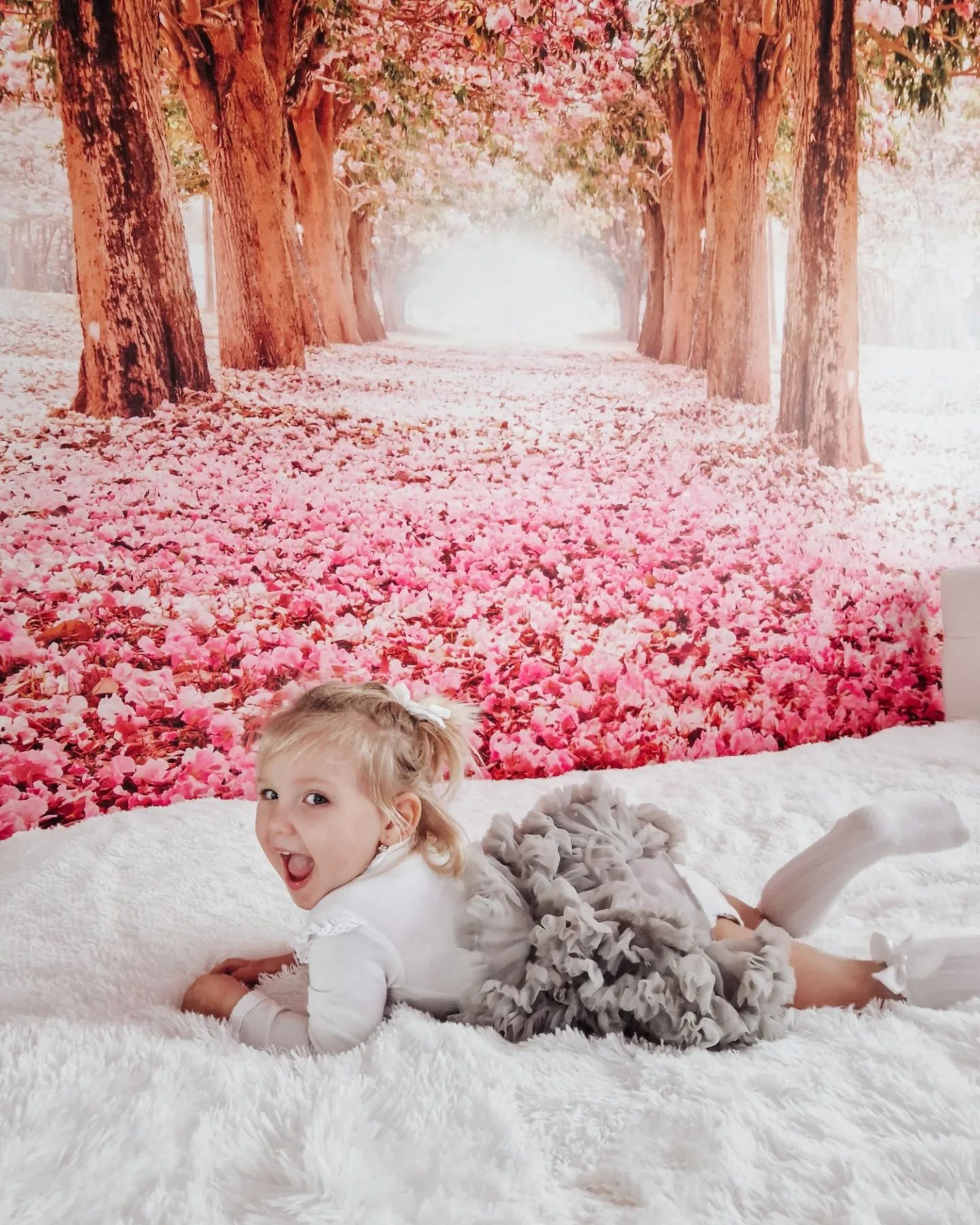 A toddler in a gray tutu lies on a white blanket with a pink blossom backdrop.
