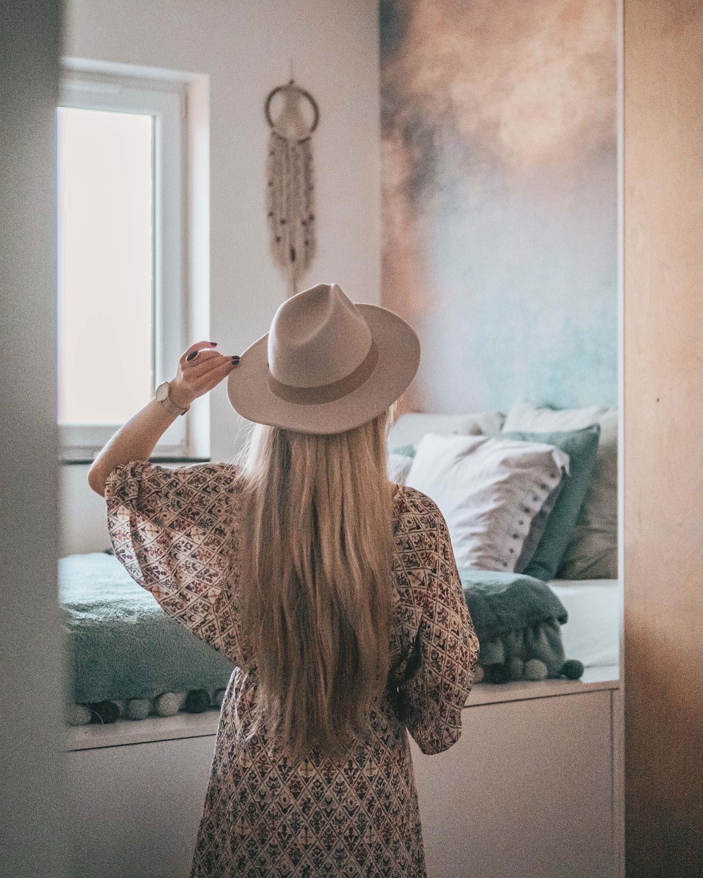 Blonde Frau in gemustertem Kleid und Hut, die einem Bett mit Kissen und einer Überwurfdecke zugewandt ist. Traumfänger an der Wand.