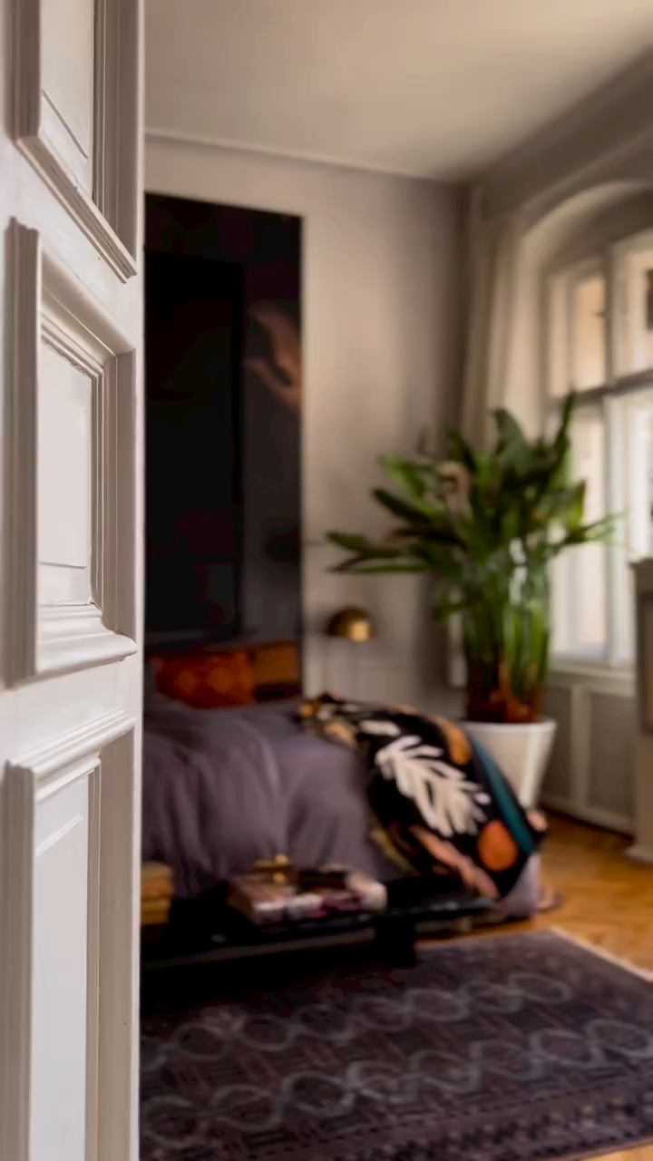 Blurred view of bedroom through white door, featuring bed, plant, and patterned rug.