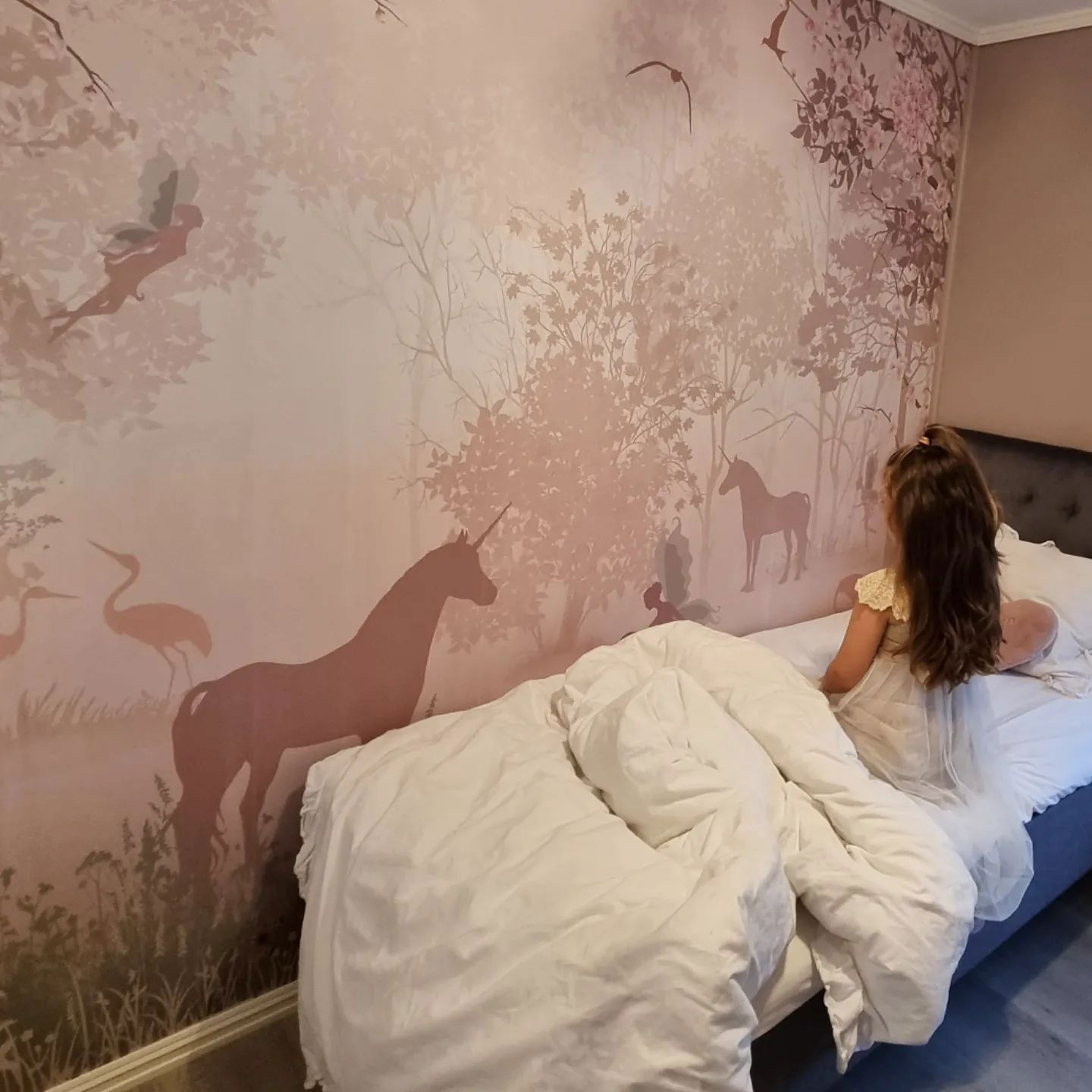 A child sits on a bed, gazing at a pink mural of unicorns, fairies, and birds.