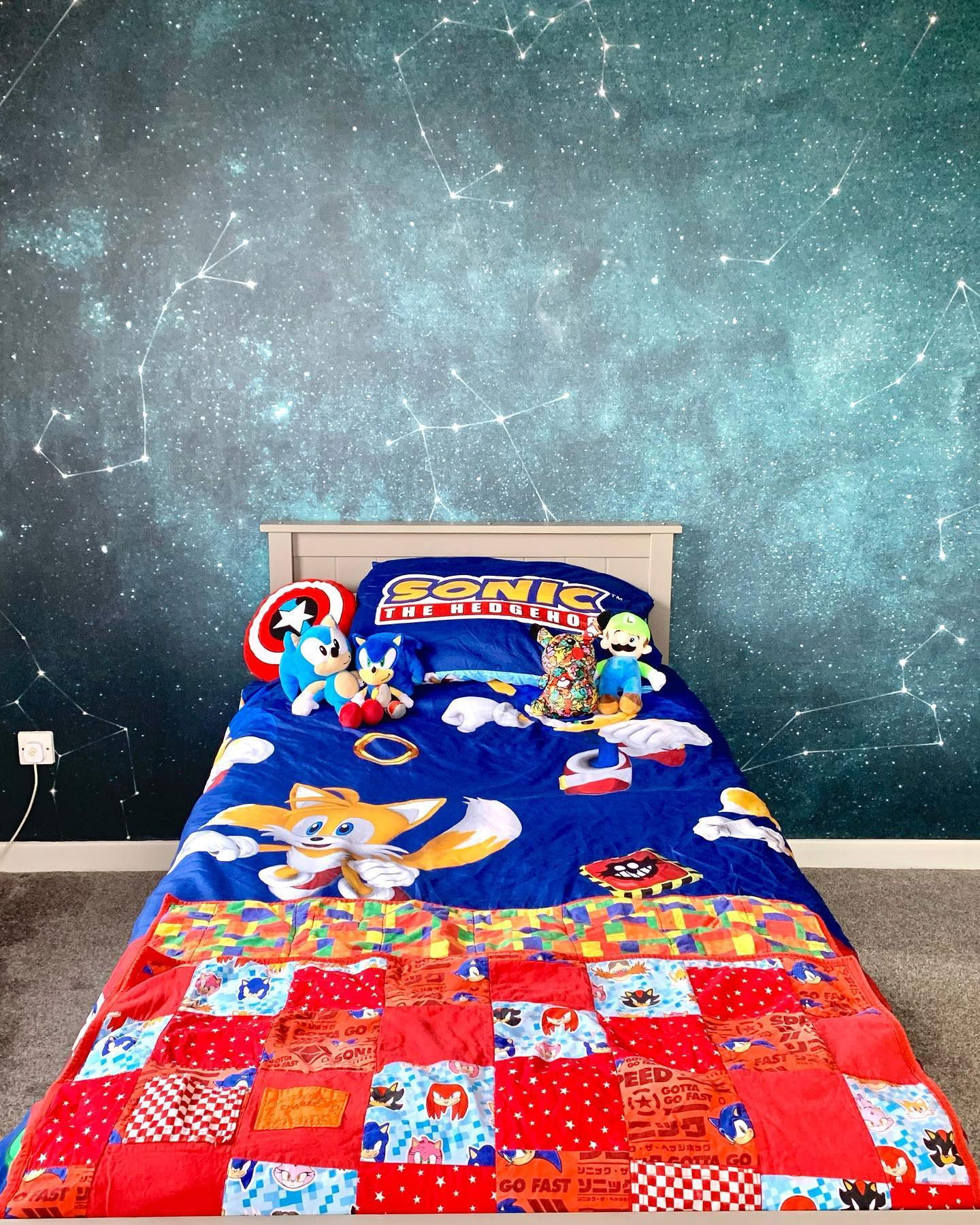 Child's bed with Sonic the Hedgehog bedding, quilt, and plush toys against a constellation wallpaper.