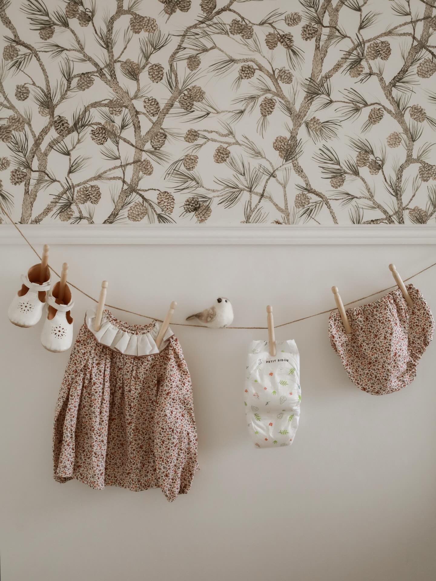 Baby clothes and a stuffed bird hang on a clothesline, with pinecone wallpaper above.
