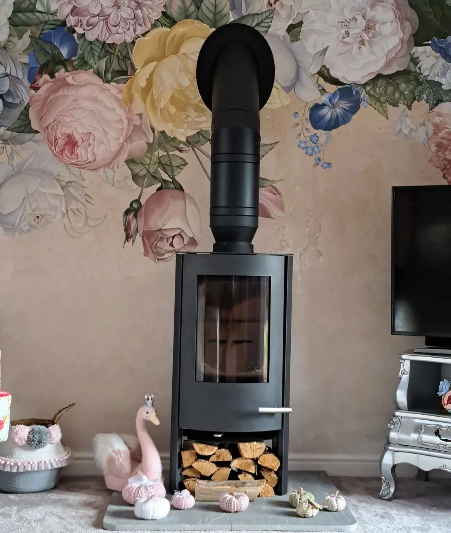Sala de estar con estufa de leña negra, papel tapiz floral y decoración de cisne rosa.