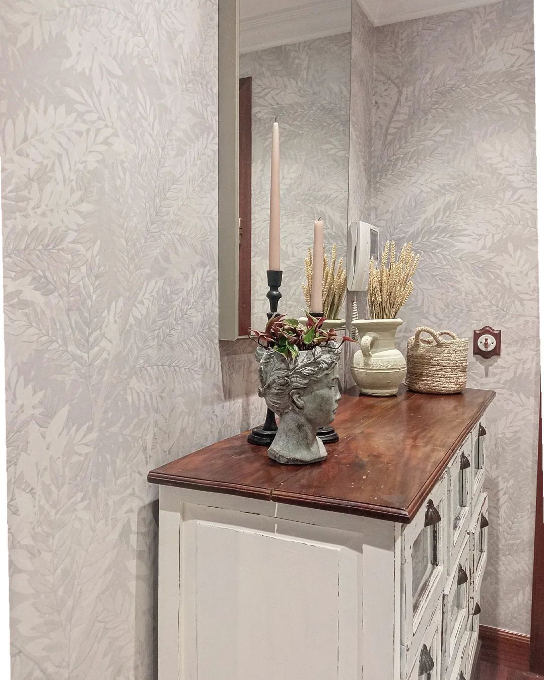 Hallway dresser with head planter, candles, wheat, and basket. Leafy wallpaper.