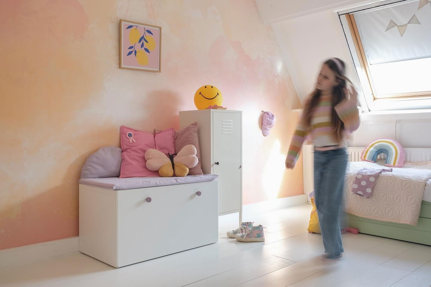 Child's bedroom with white storage bench, pillows, locker, and bed. Girl walks by.