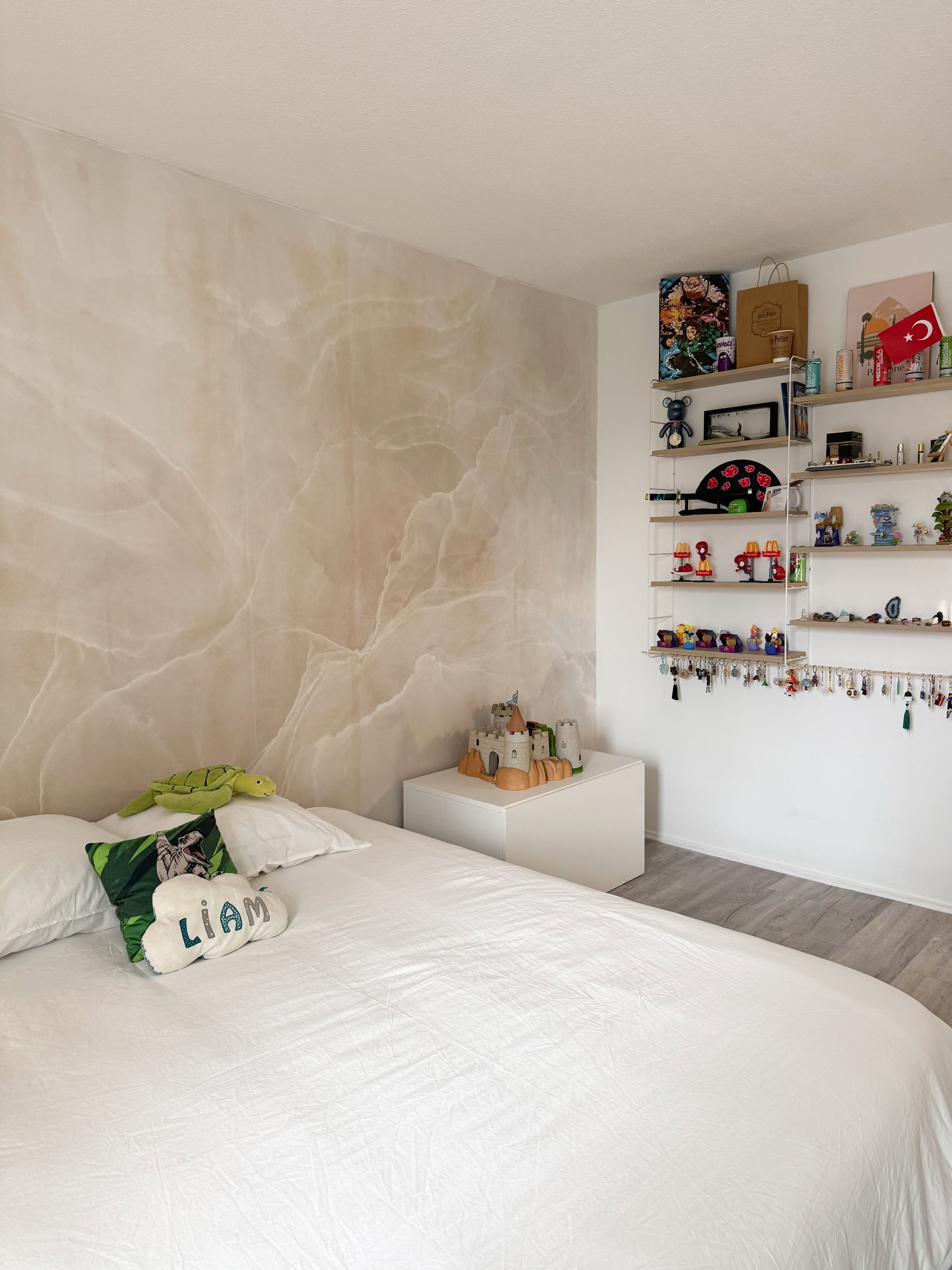Dormitorio con una cama blanca, una pared de mármol beige y estanterías blancas que exhiben objetos. Una tortuga de peluche verde y una almohada con 'LIAM' están en la cama.