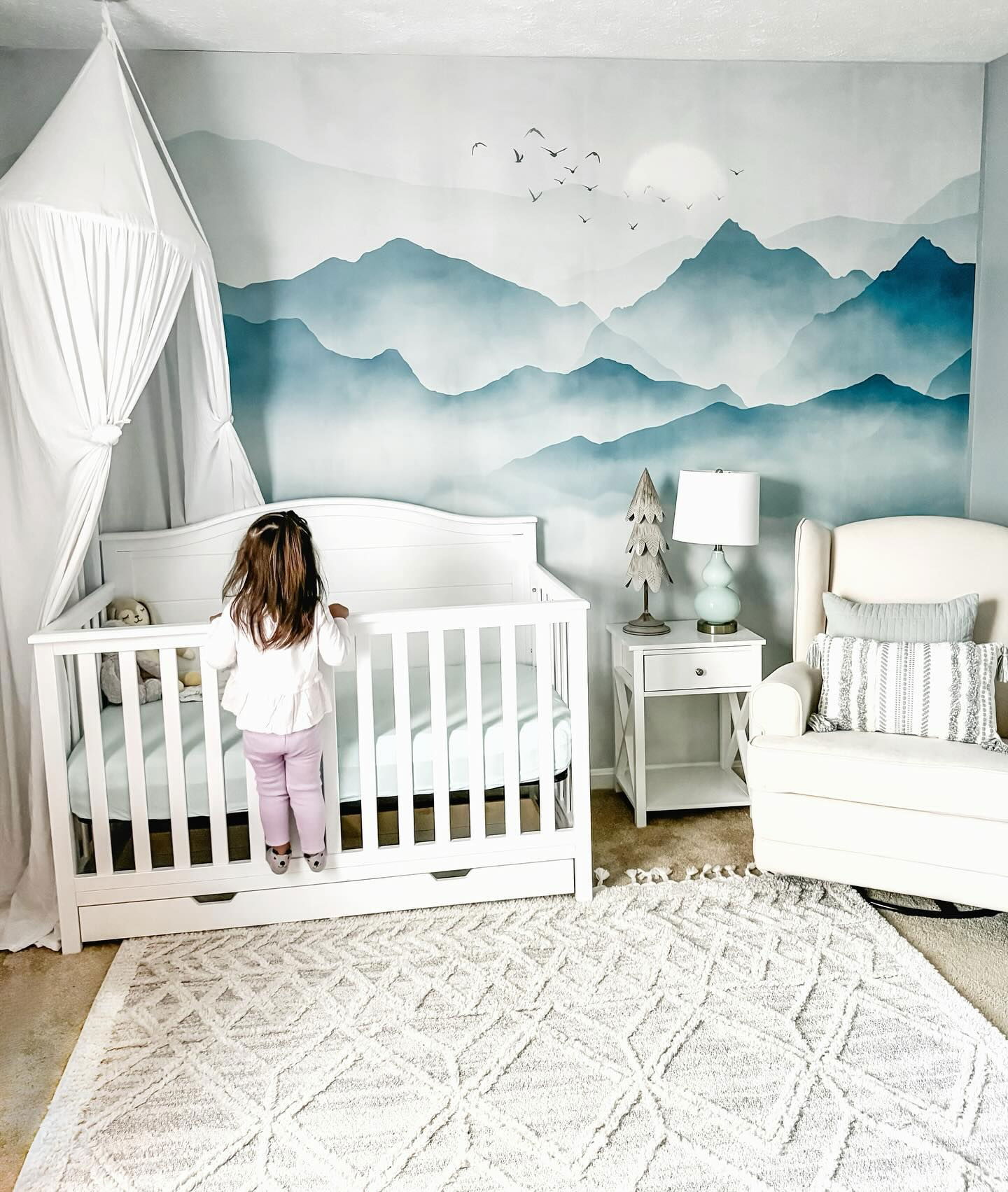 Nursery with white crib, mountain mural, and patterned rug. Child stands in crib.