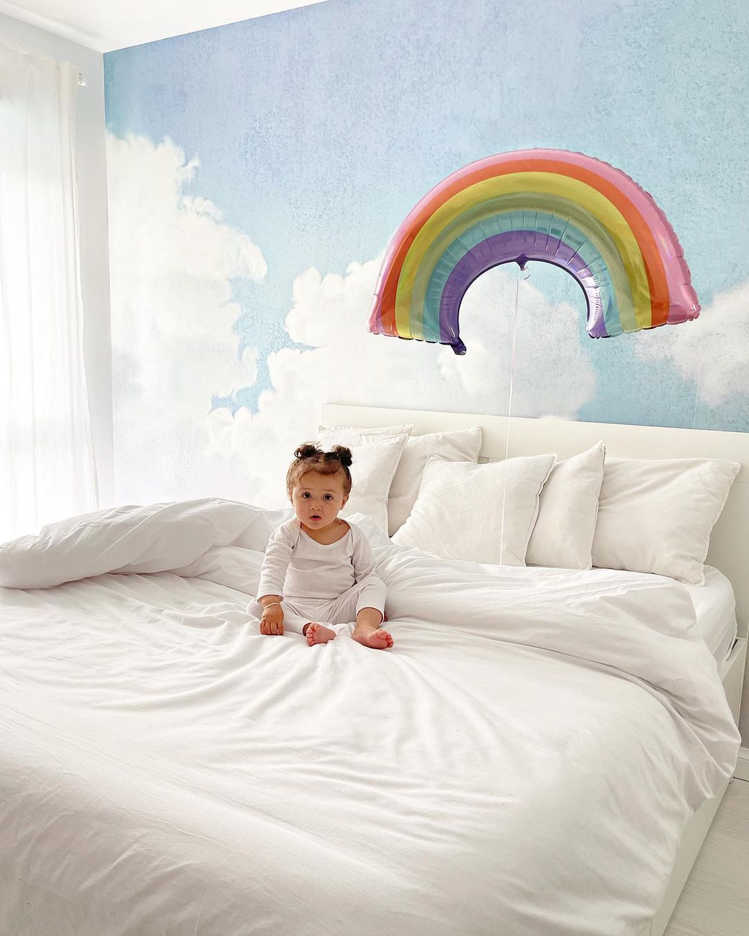A baby sits on a white bed with a rainbow balloon and cloud wallpaper.