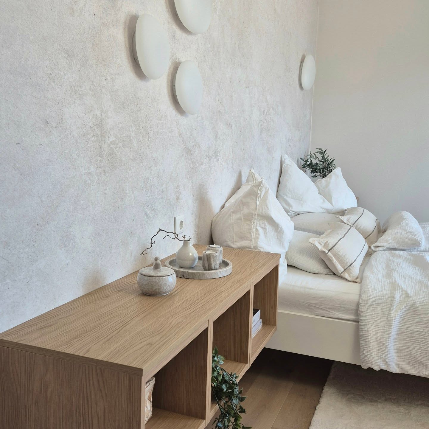Minimalistyczna sypialnia z szafką nocną z jasnego drewna, białą pościelą i teksturowanymi ścianami.