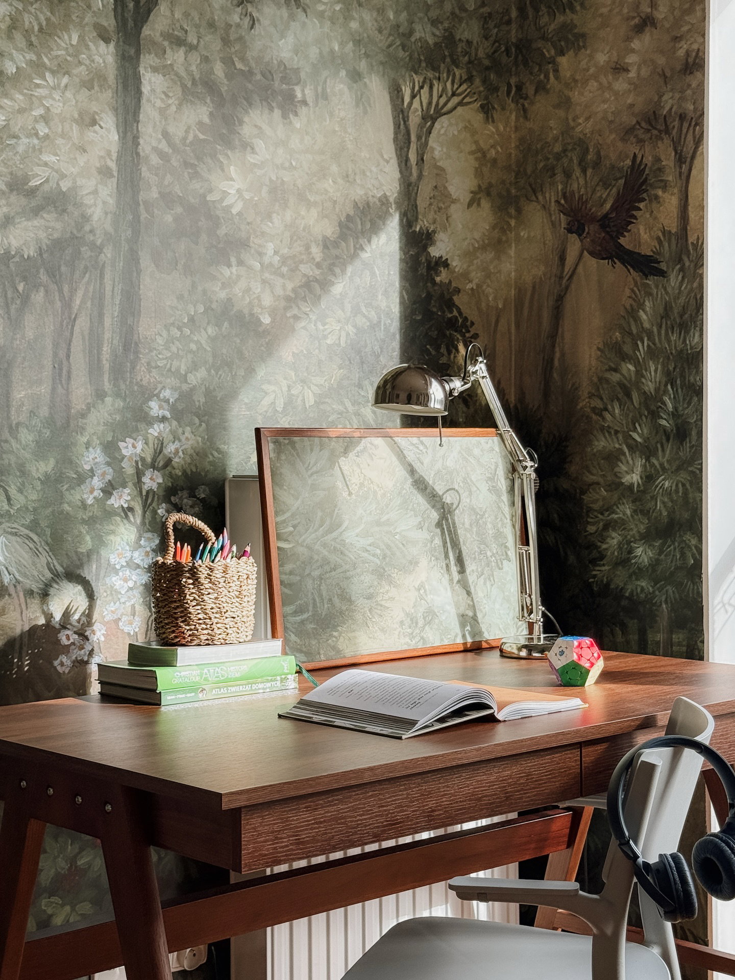 Bruin houten bureau met een open boek, groene boeken, mand met potloden, zilveren lamp en geometrisch puzzel, tegen een bosbehang met zonlicht.