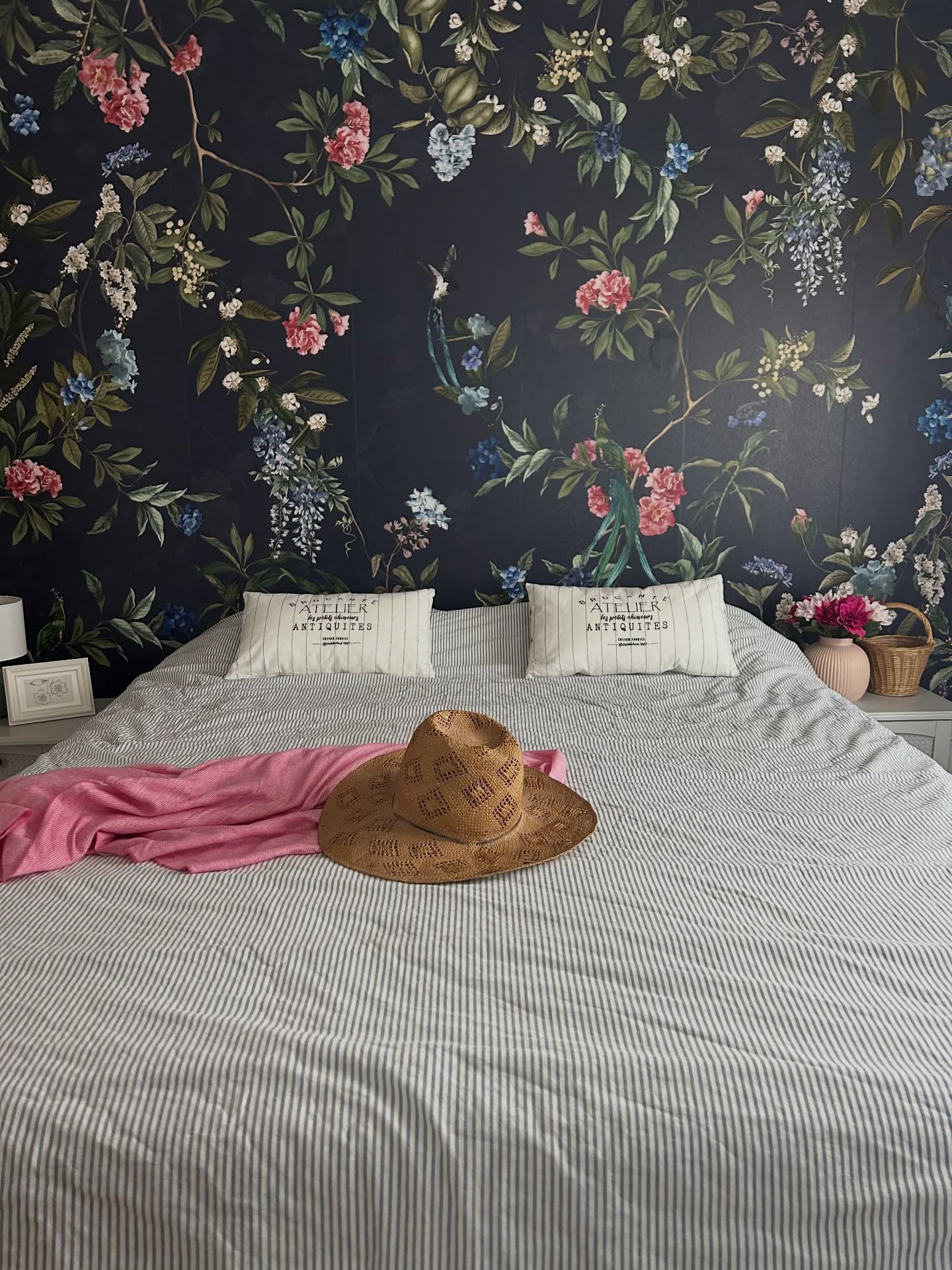Bed with floral wallpaper, striped bedding, a straw hat, and a pink throw.