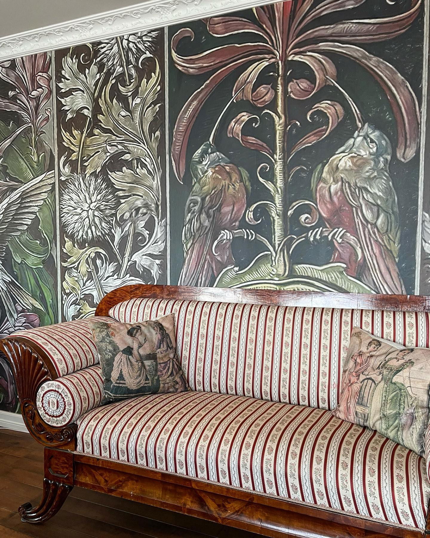 Antik randig soffa med dekorativa kuddar mot en blommig väggmålning.