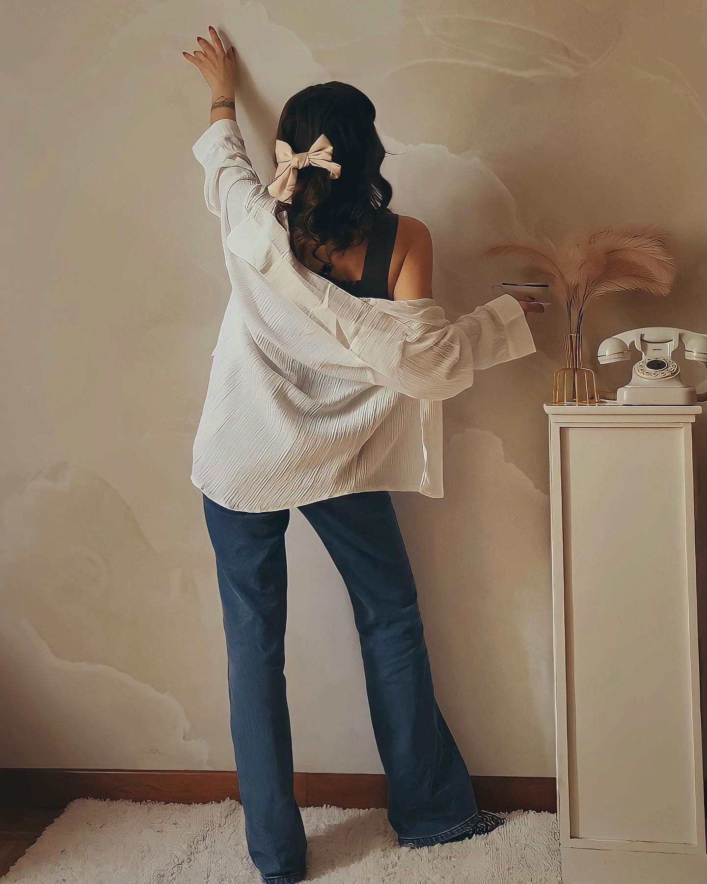 Vista posteriore di donna in jeans, camicia bianca e fiocco per capelli. Telefono retrò e vaso sull'armadio.
