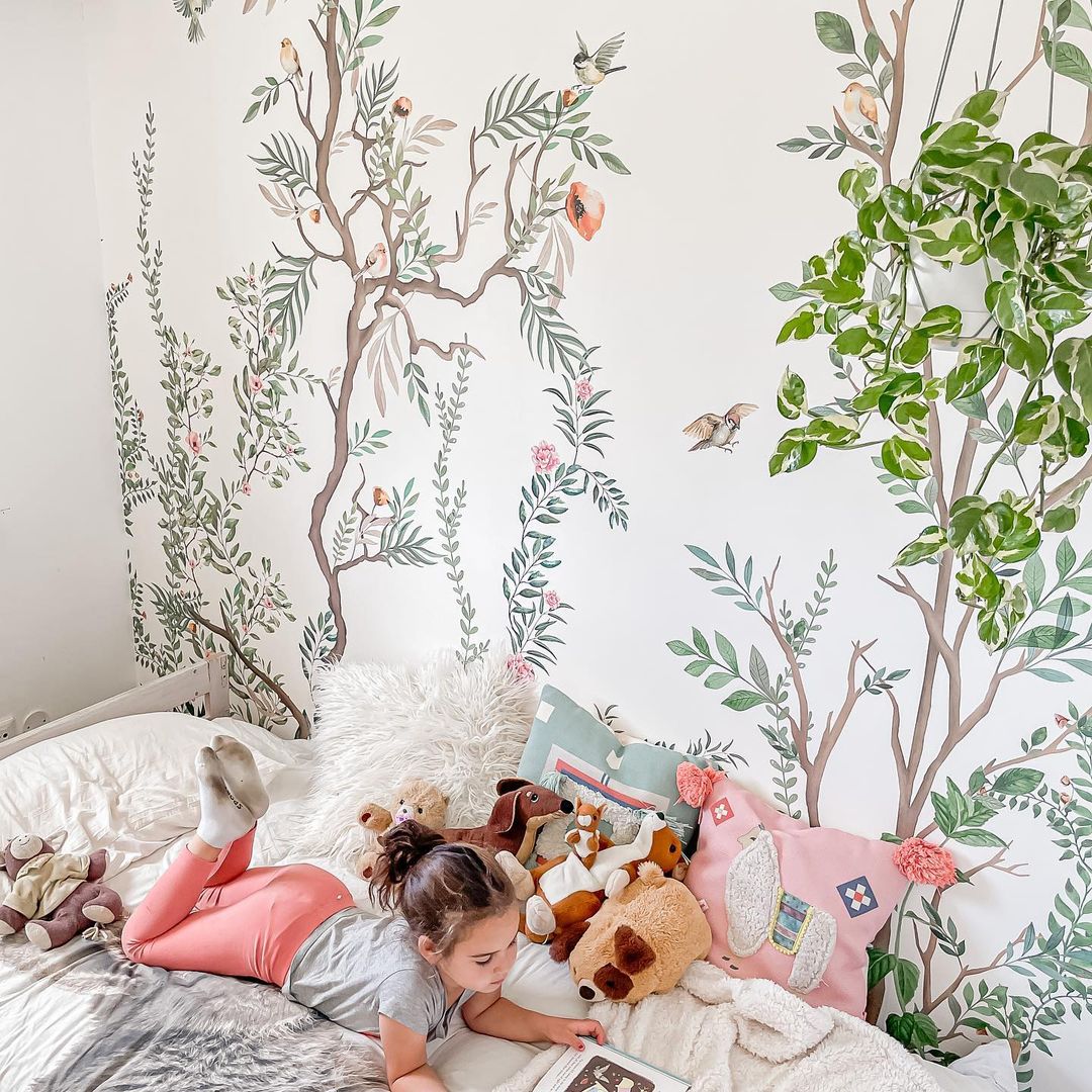 A young girl reads on a bed with stuffed animals and decorative pillows, set against a floral mural.