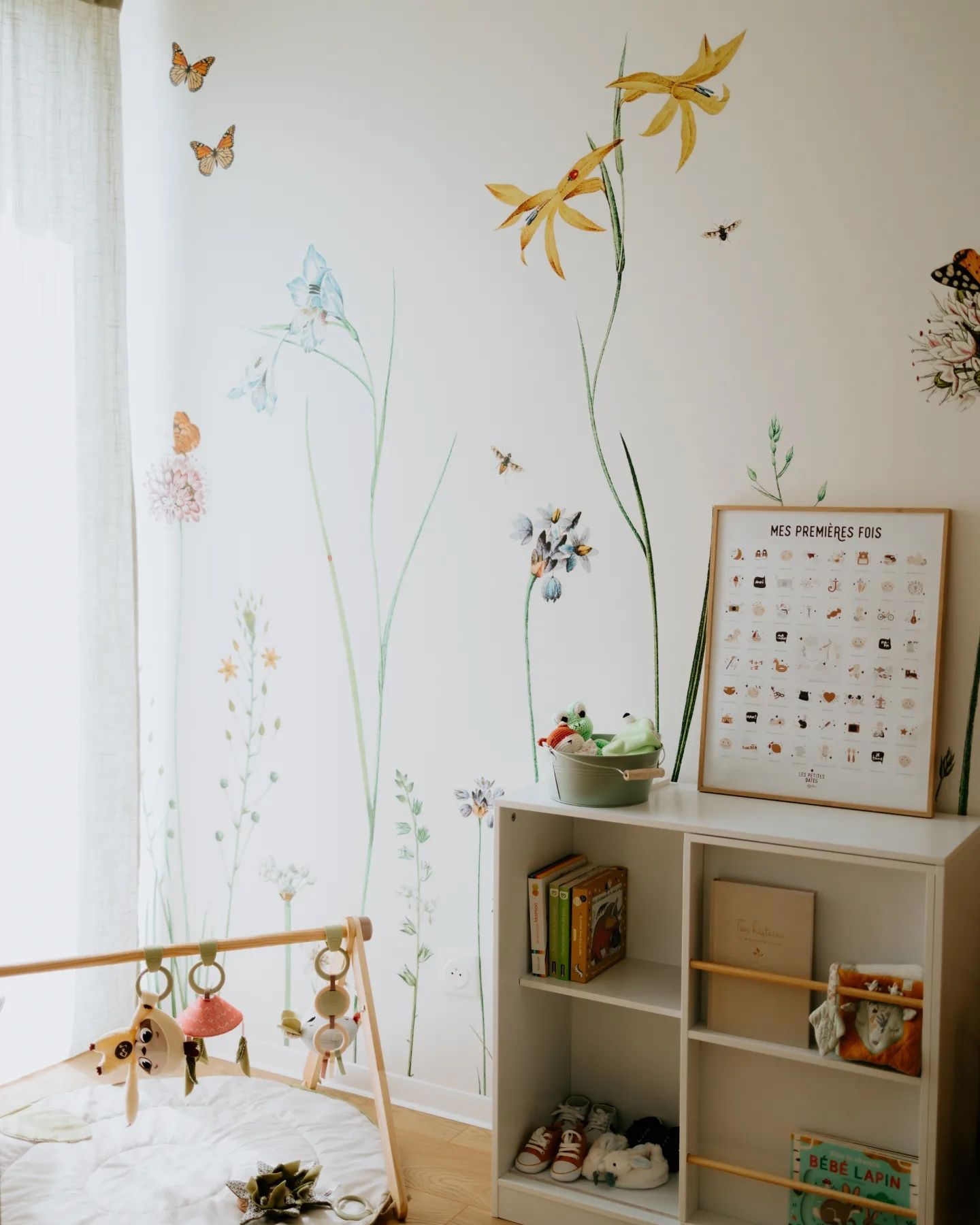 Chambre de bébé avec décorations murales florales, tapis d'éveil et étagère blanche remplie de livres et de jouets.