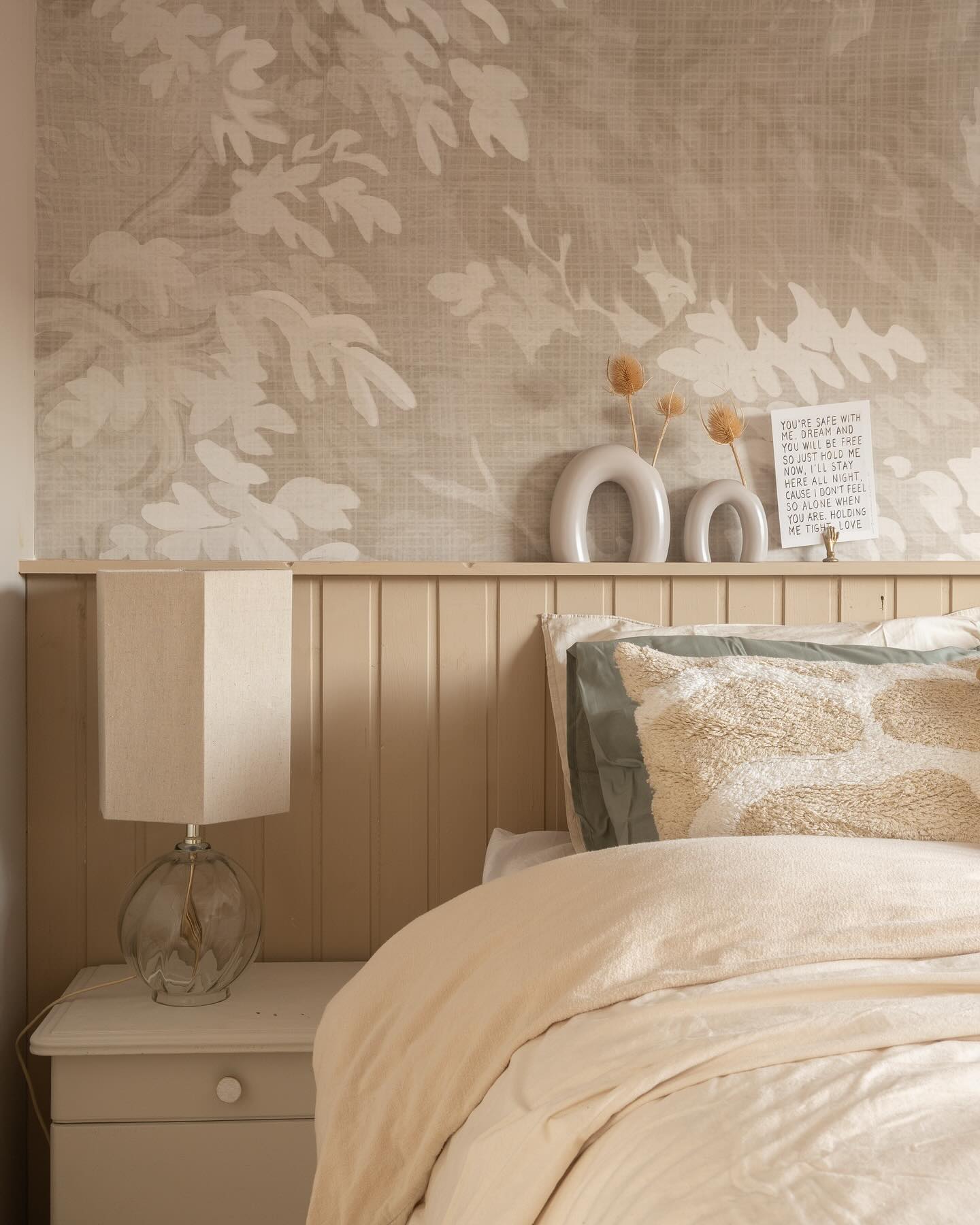 Bedroom with beige floral wallpaper, lamp on nightstand, and bed with neutral bedding.