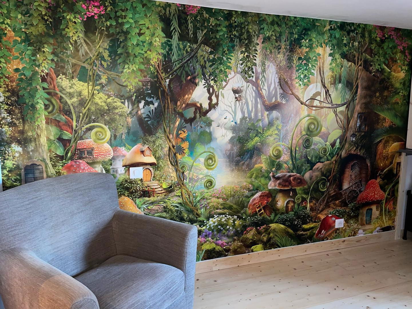 Living room with gray armchair and fairy tale forest mural featuring mushroom houses.