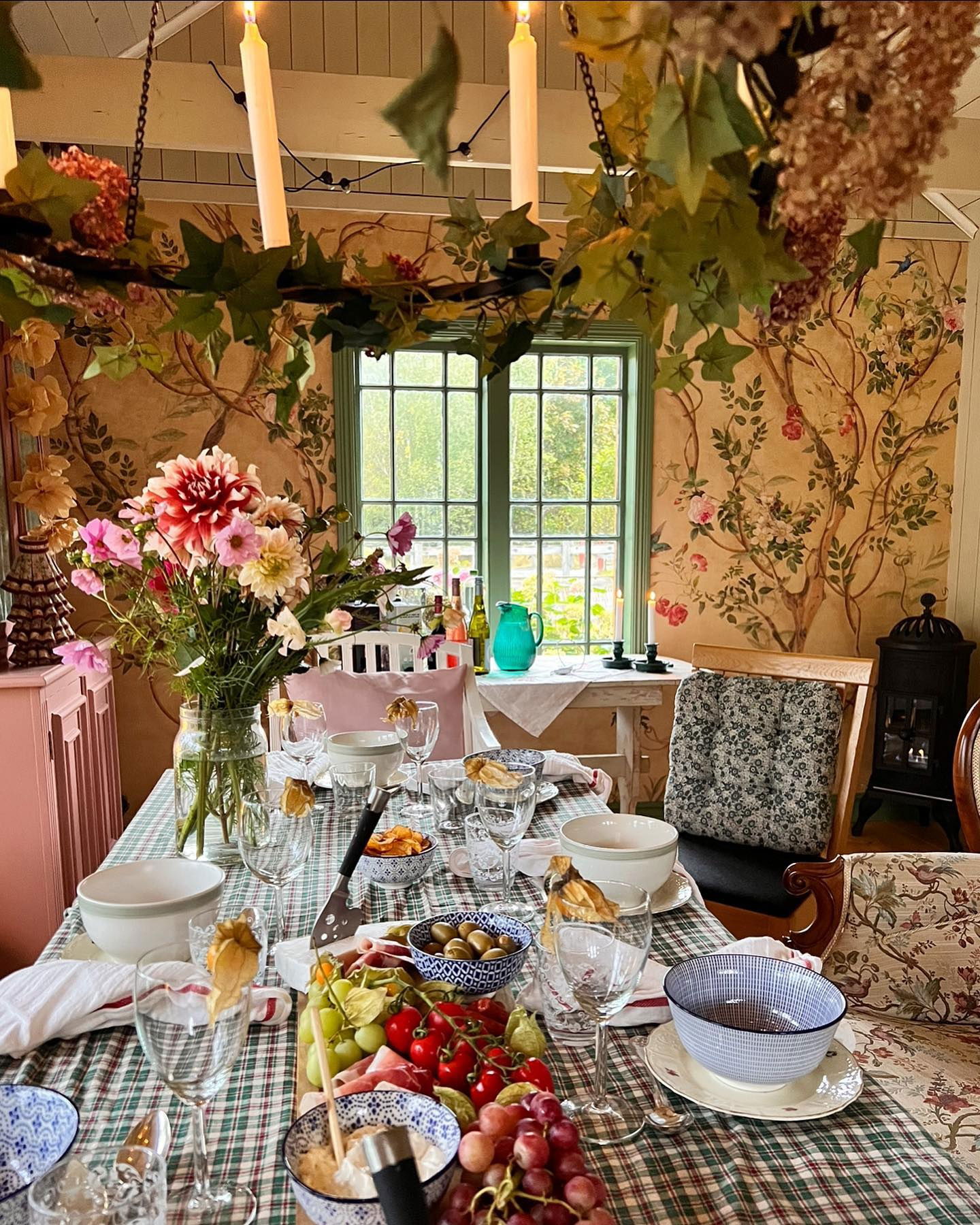 Festligt spisebord dækket med blomster, frugter og lys i et rum med blomstret tapet.