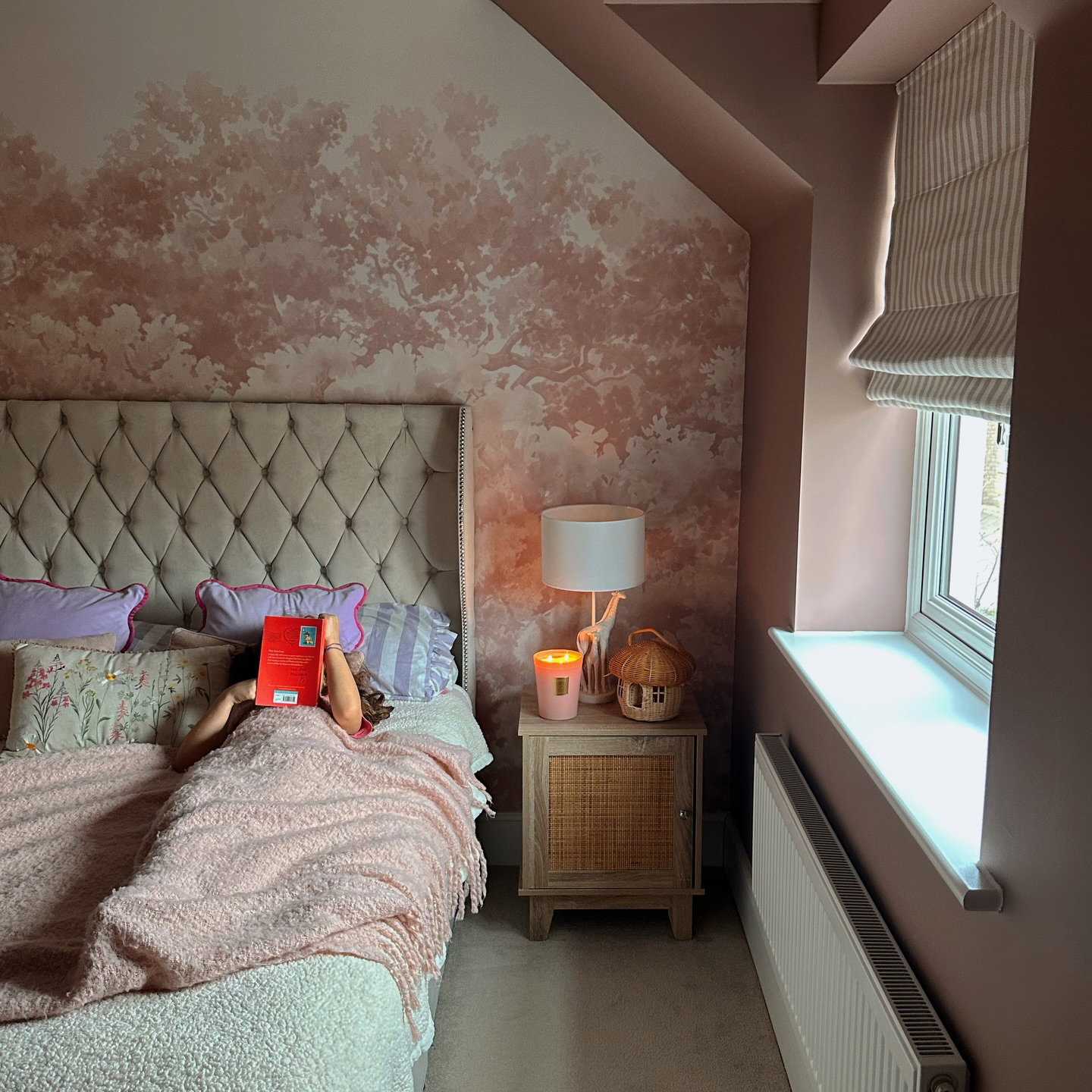 Una persona debajo de una manta rosa lee un libro rojo en la cama. La habitación rosa tiene una pared con estampado de árboles y una mesita de noche con una lámpara encendida.