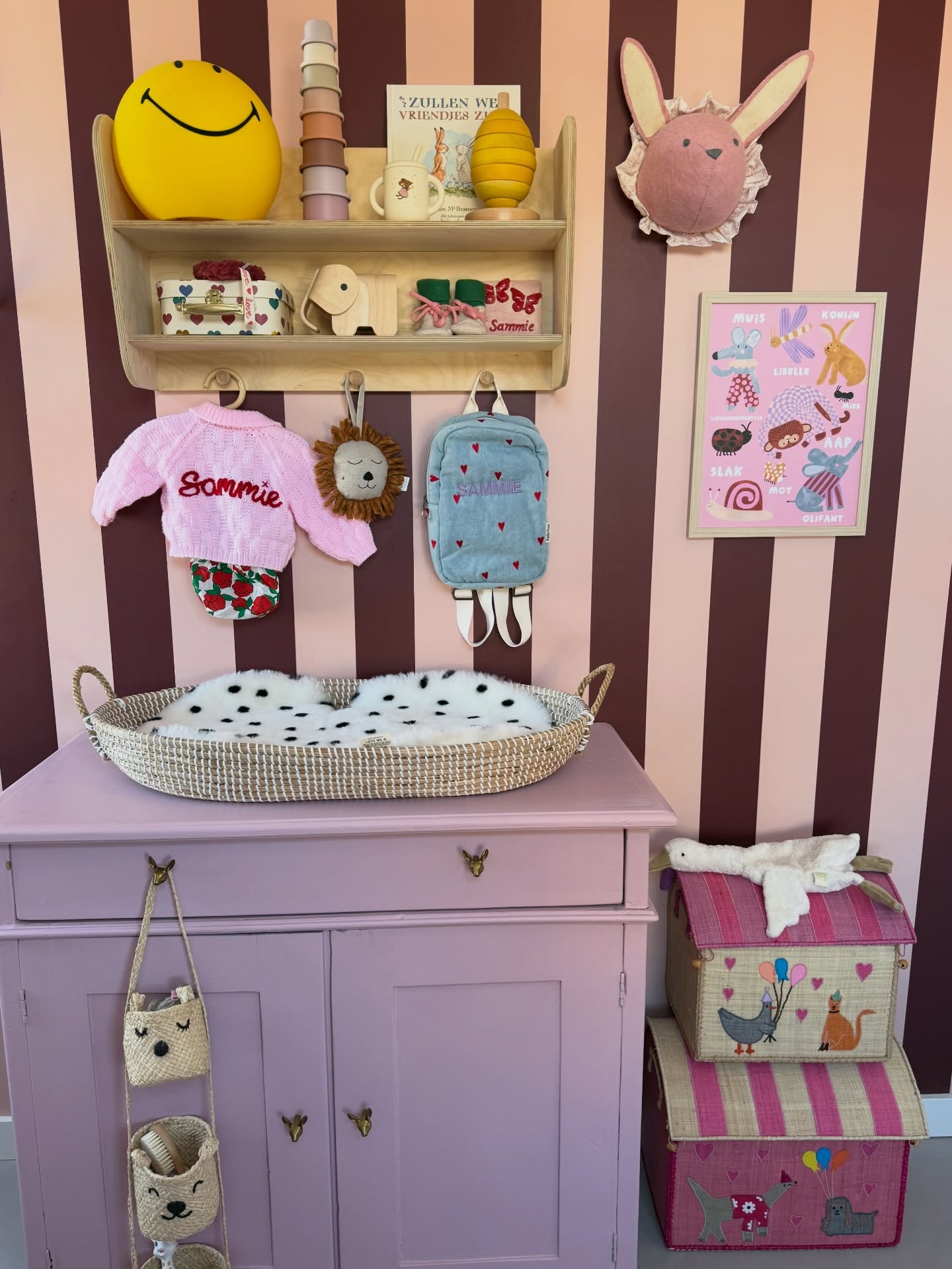 Pink dresser with a woven basket, baby clothes, and toys on striped walls.