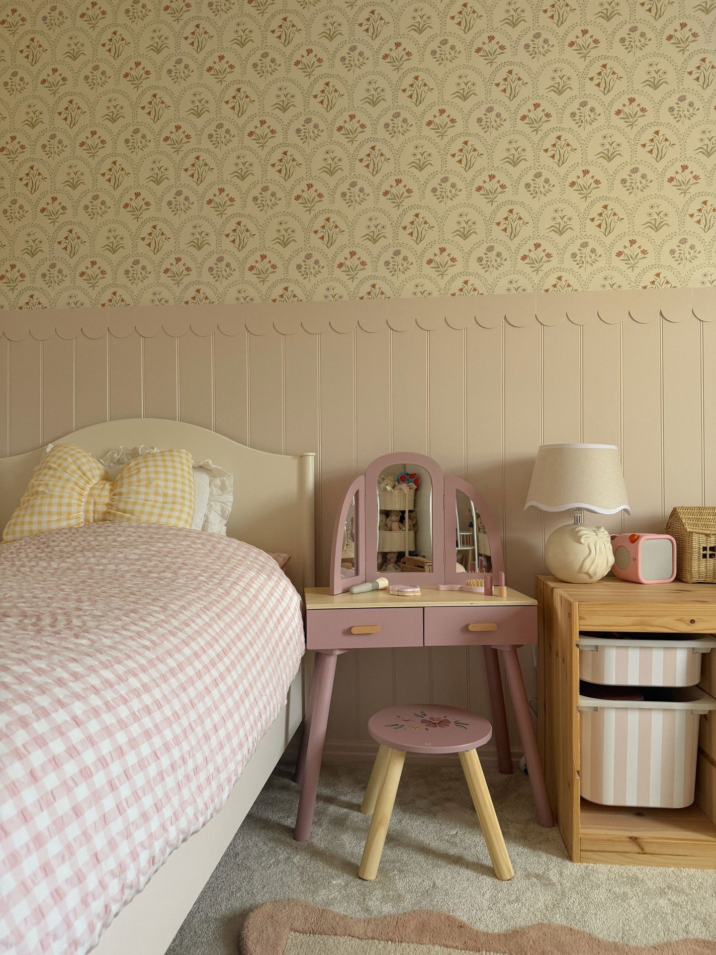 Cama de cuadros rosa, tocador rosa con espejo, estante de madera con contenedores de almacenamiento y papel tapiz floral.