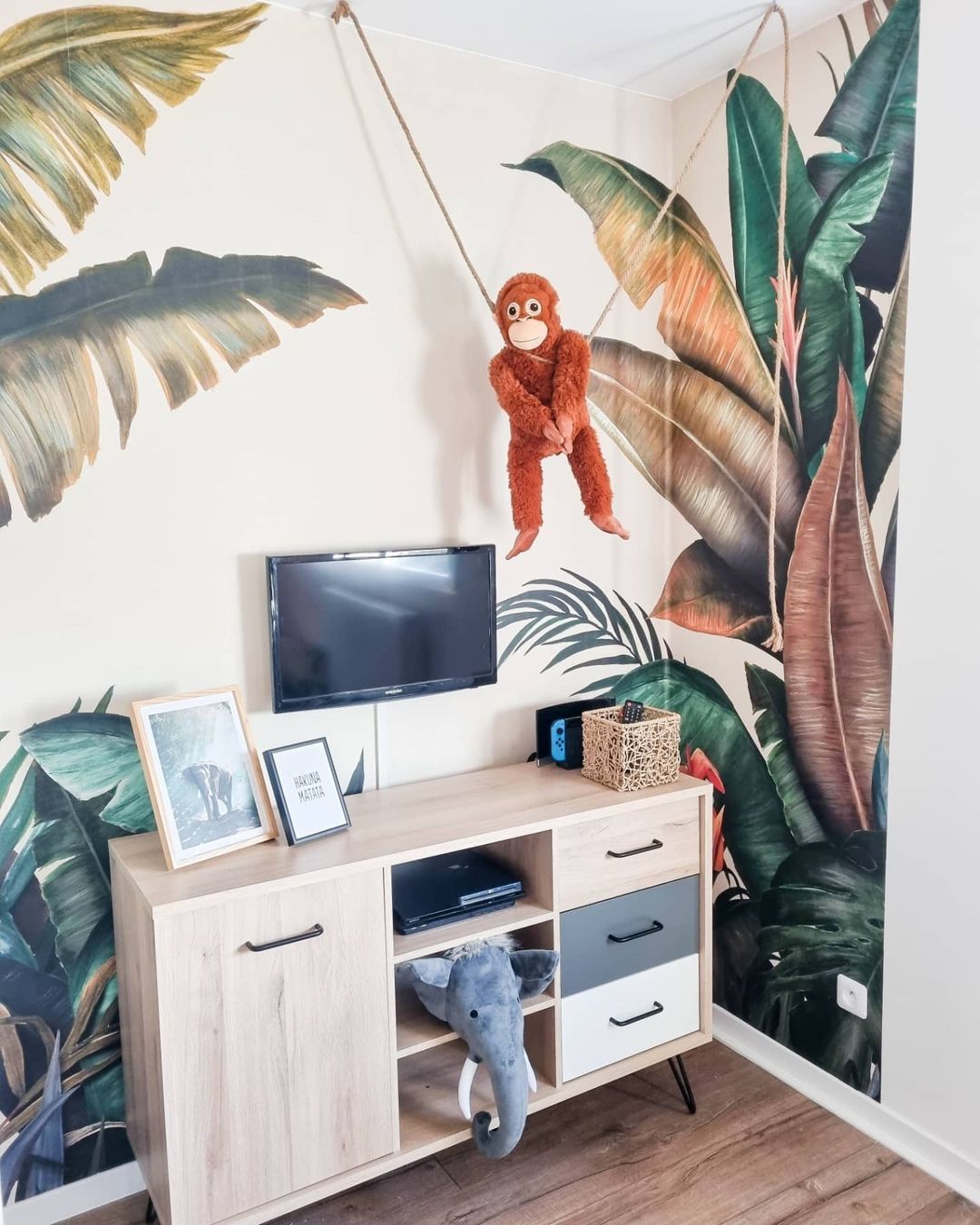 Child's room with jungle wallpaper, TV, and light wood cabinet. A stuffed monkey hangs from the ceiling.