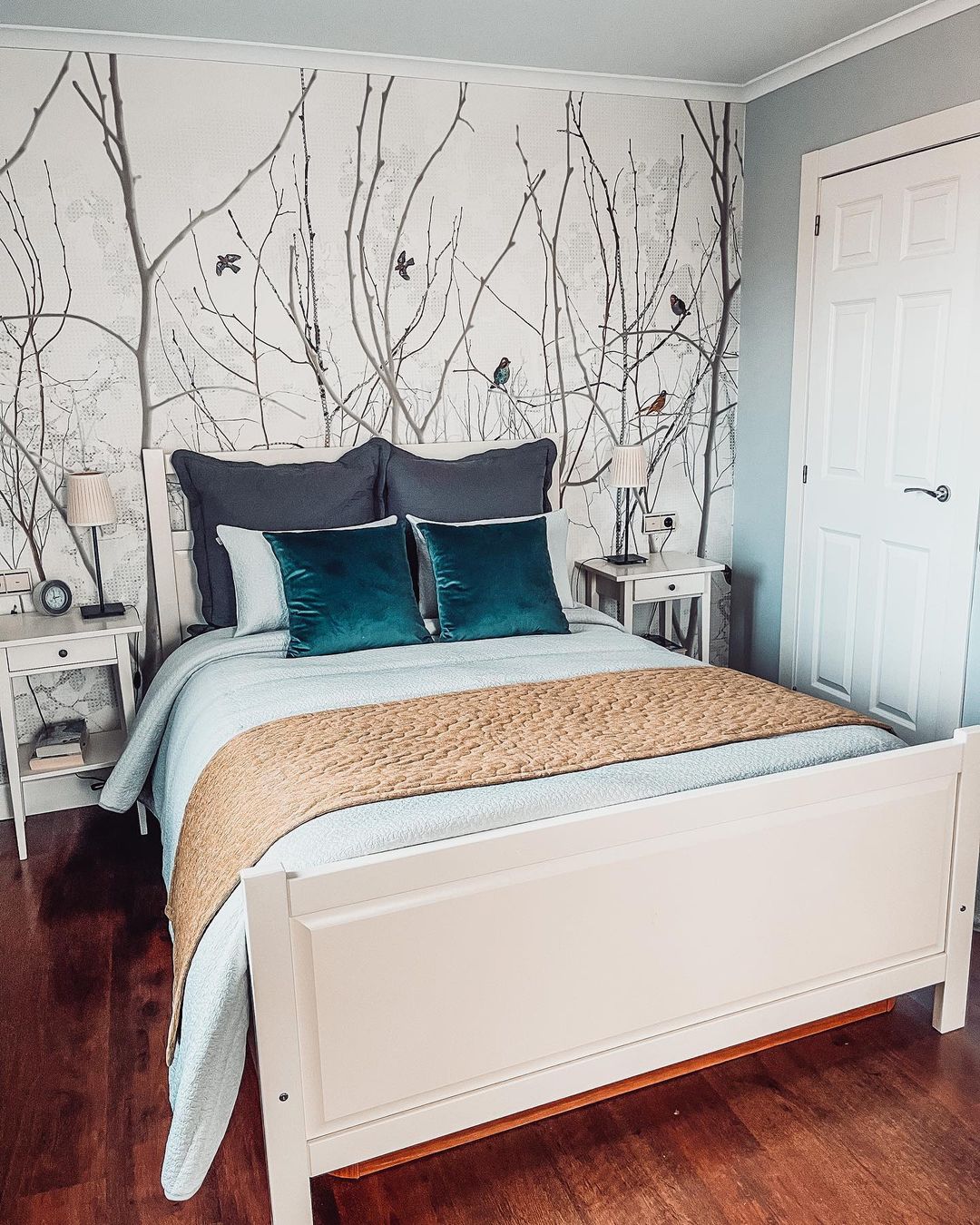 Bedroom with white bed, teal pillows, and tree-and-bird wallpaper.