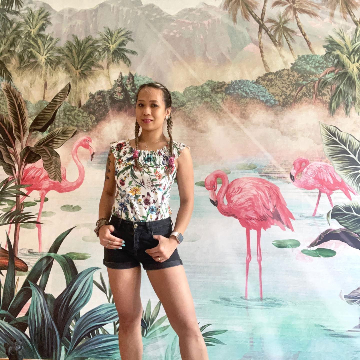 En kvinde i en blomstret top og shorts poserer foran et flamingo- og palmetræsmaleri.