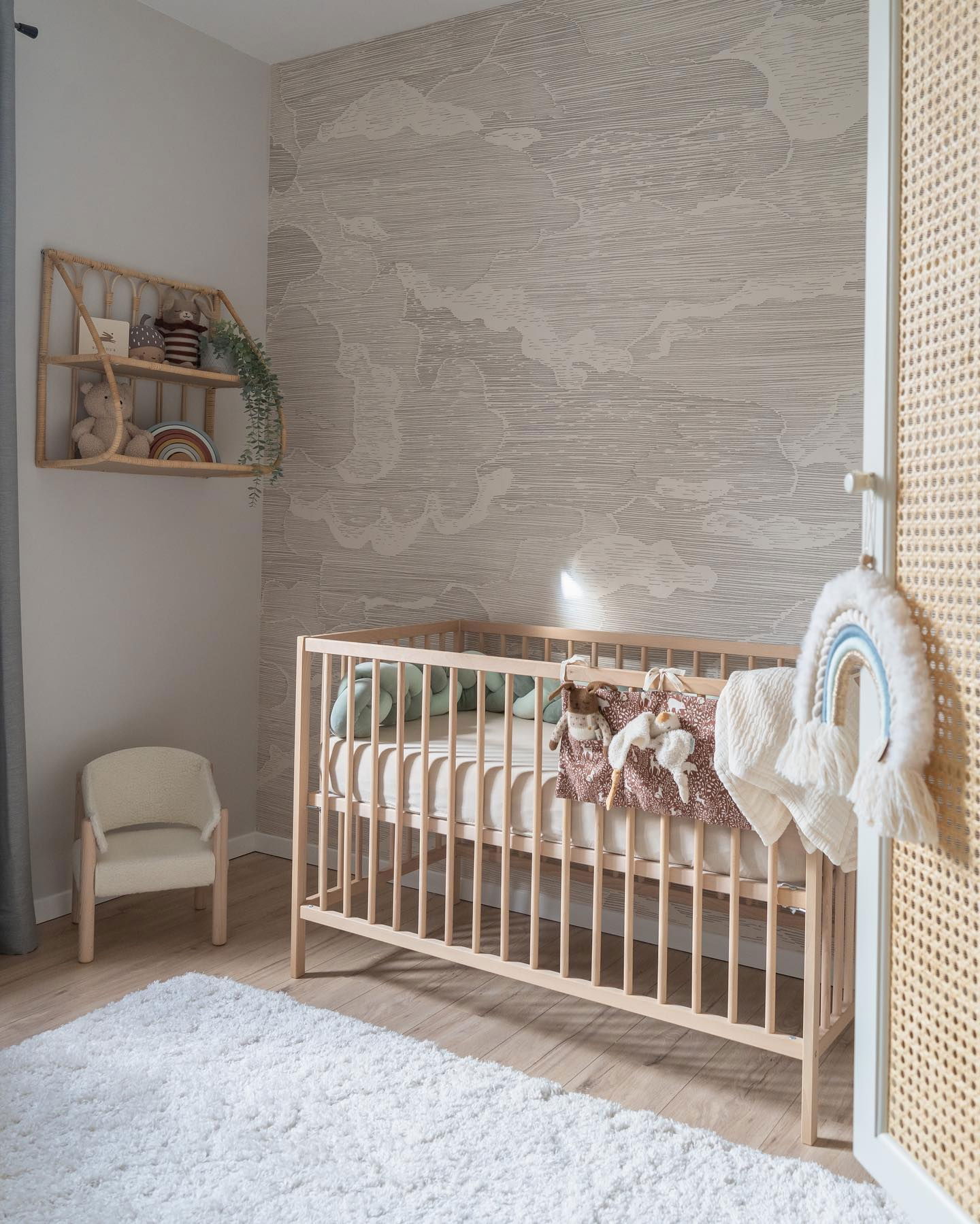 Nursery with wooden crib, cloud wallpaper, and white shag rug. Rattan shelf with toys and rainbow decor.