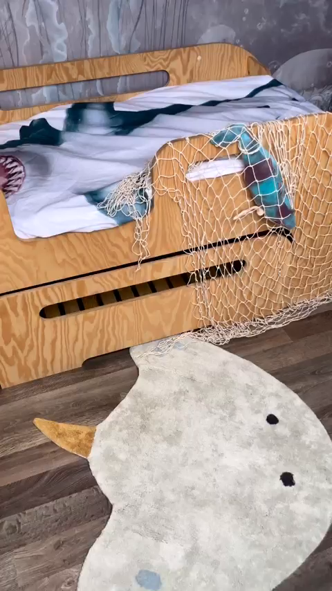 Nautical themed child's room with wooden bed, shark bedding, fishnet decor, and narwhal shaped rug.