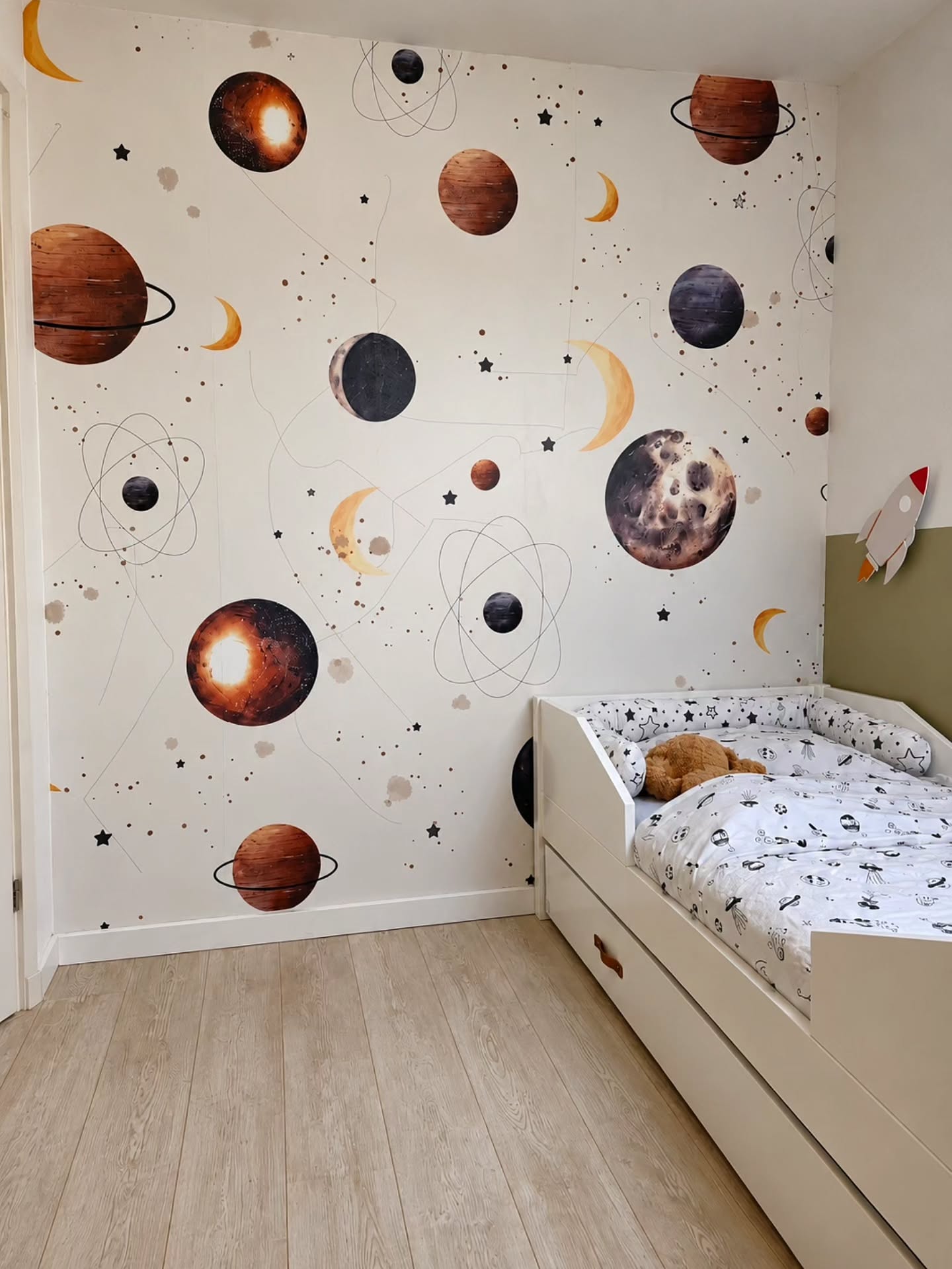 Ein Kinderzimmer mit Weltraum-Tapeten, die Planeten und Monde zeigen. Ein weißes Bett mit Schubladen und gemusterter Bettwäsche steht auf einem hellen Holzboden. Ein Teddybär liegt auf dem Bett.