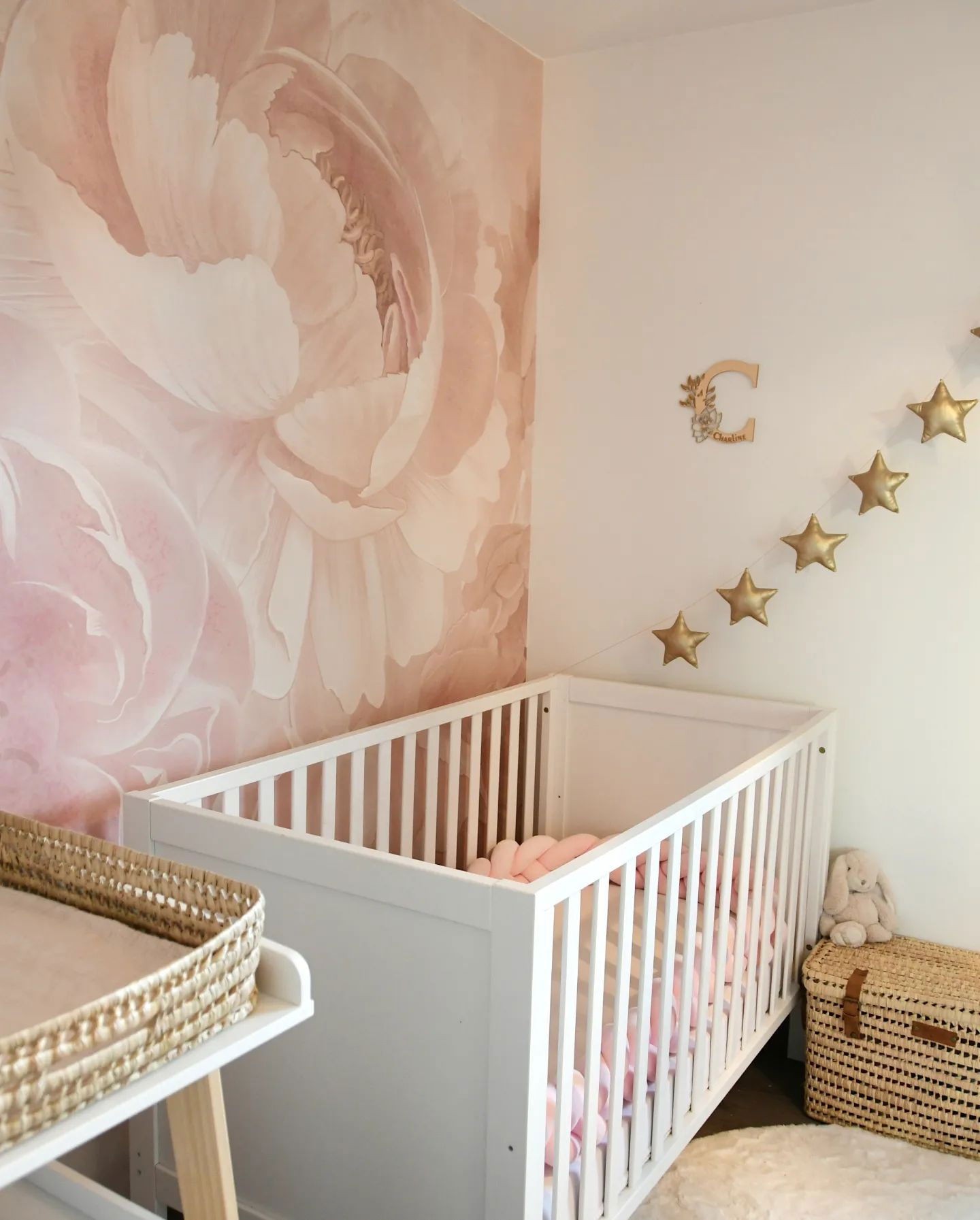 Stanza dei bambini con culla bianca, carta da parati floreale rosa e ghirlanda di stelle dorate. Cesto fasciatoio in vimini e cassapanca.