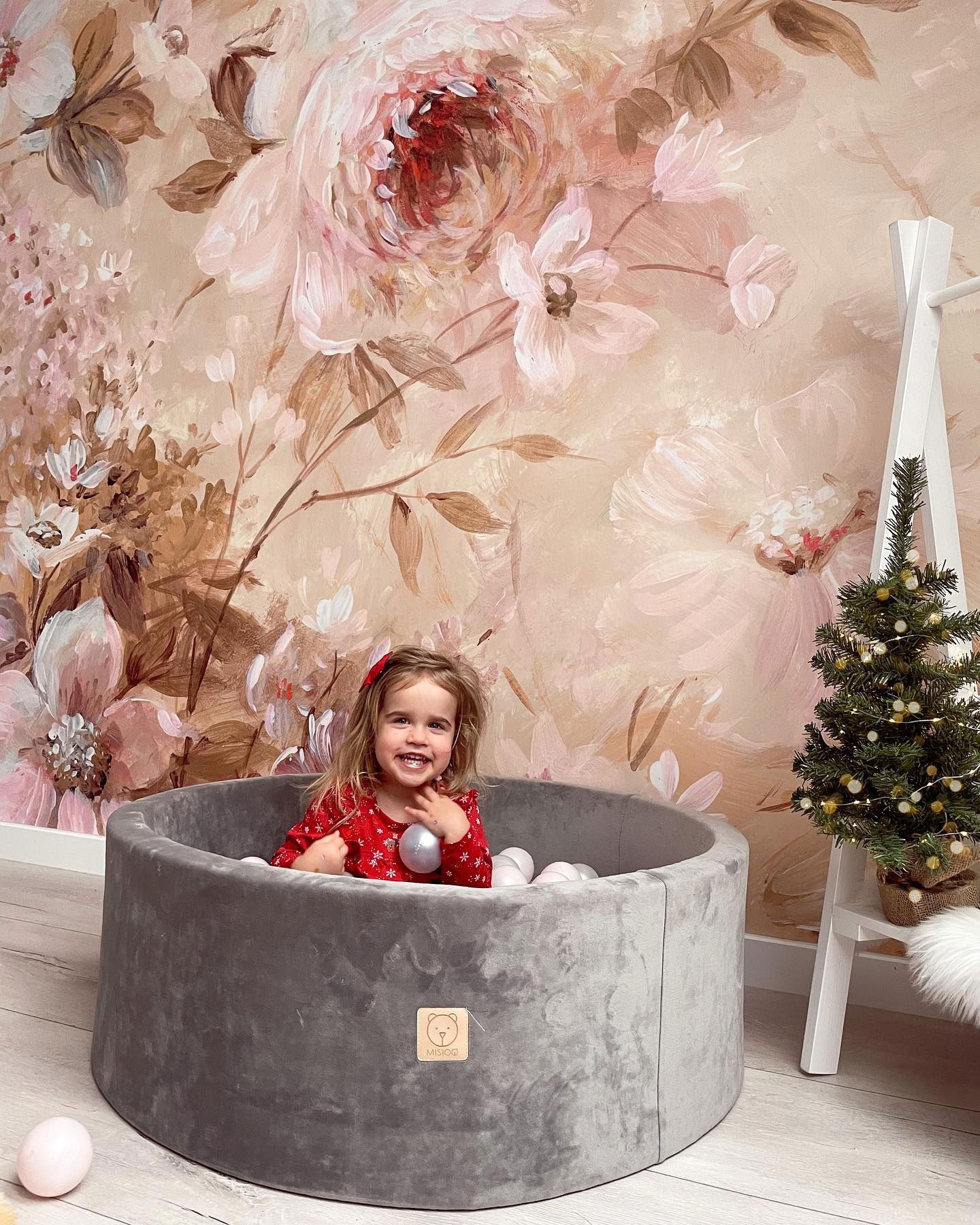 A smiling toddler in a Misioo grey velvet ball pit with a floral wall and small Christmas tree.