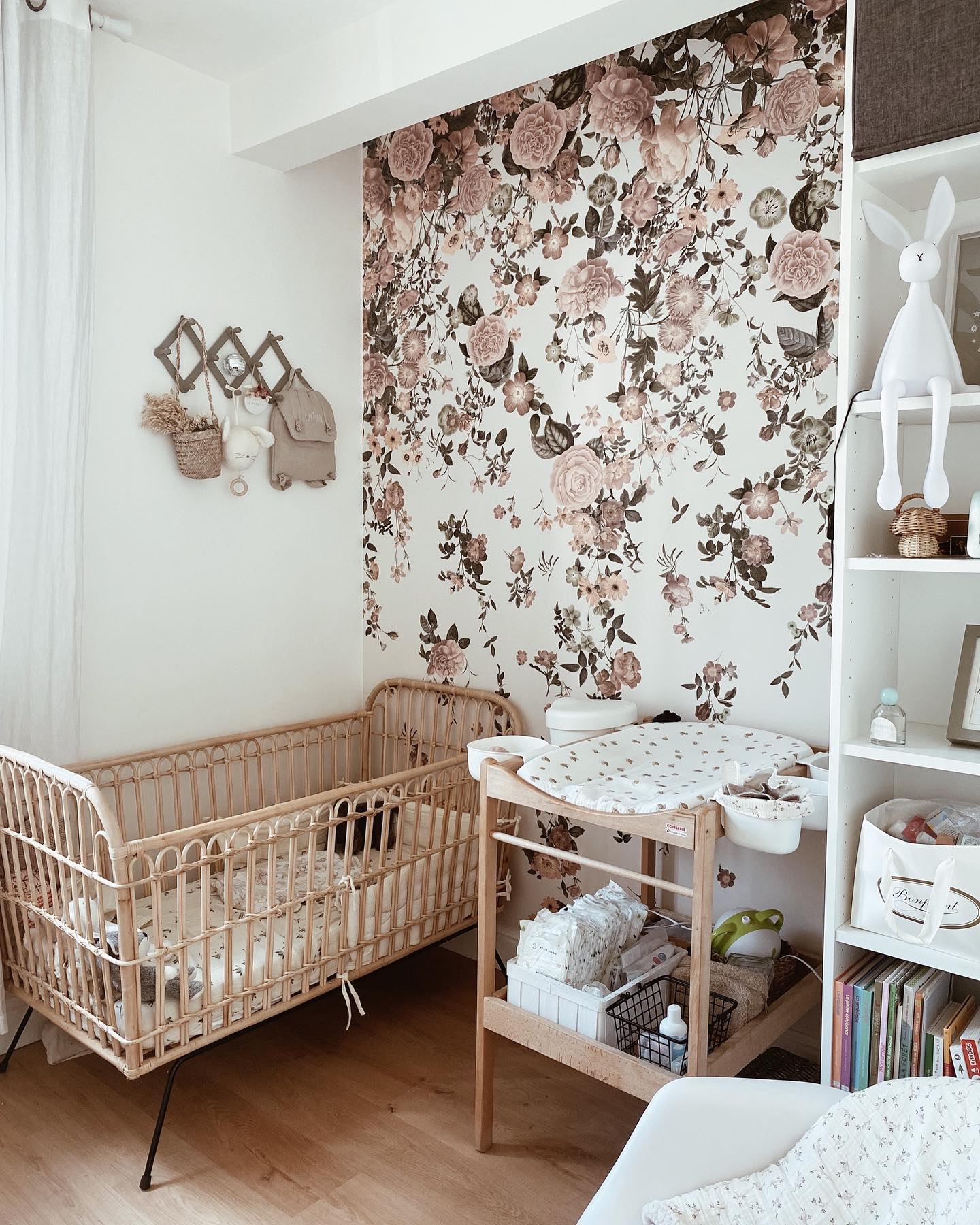 Nursery with flower wallpaper, wood crib, changing table, and white shelving.